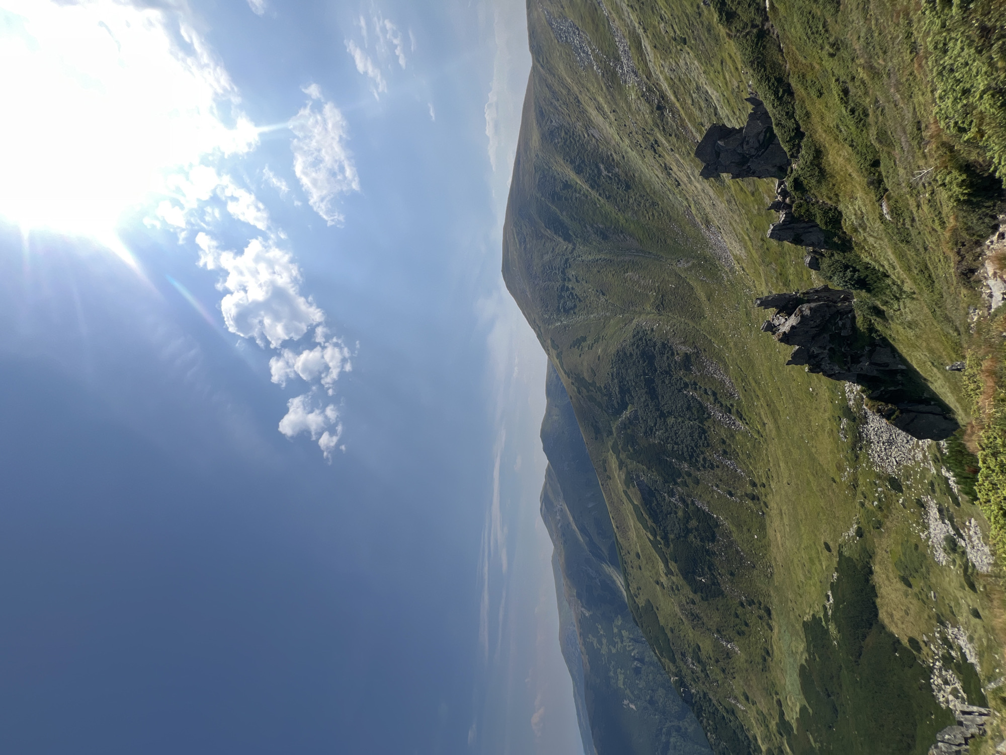This is a landscape photograph taken in the Carpathian Mountains near Vorokhta, Ukraine. The image captures rolling green hills and mountains under a bright blue sky with scattered white clouds. The sun is shining intensely, creating lens flare and casting light across the scene. In the foreground, there are large, dark rock formations on a grassy slope, with some sparse green vegetation. The middle ground shows more hills covered in grass and low-lying bushes, with darker patches indicating denser vegetation or shadows. In the background, a series of mountain ranges recede into the distance, their peaks softened by atmospheric haze. The overall impression is one of natural beauty and a vast, open mountainous landscape.