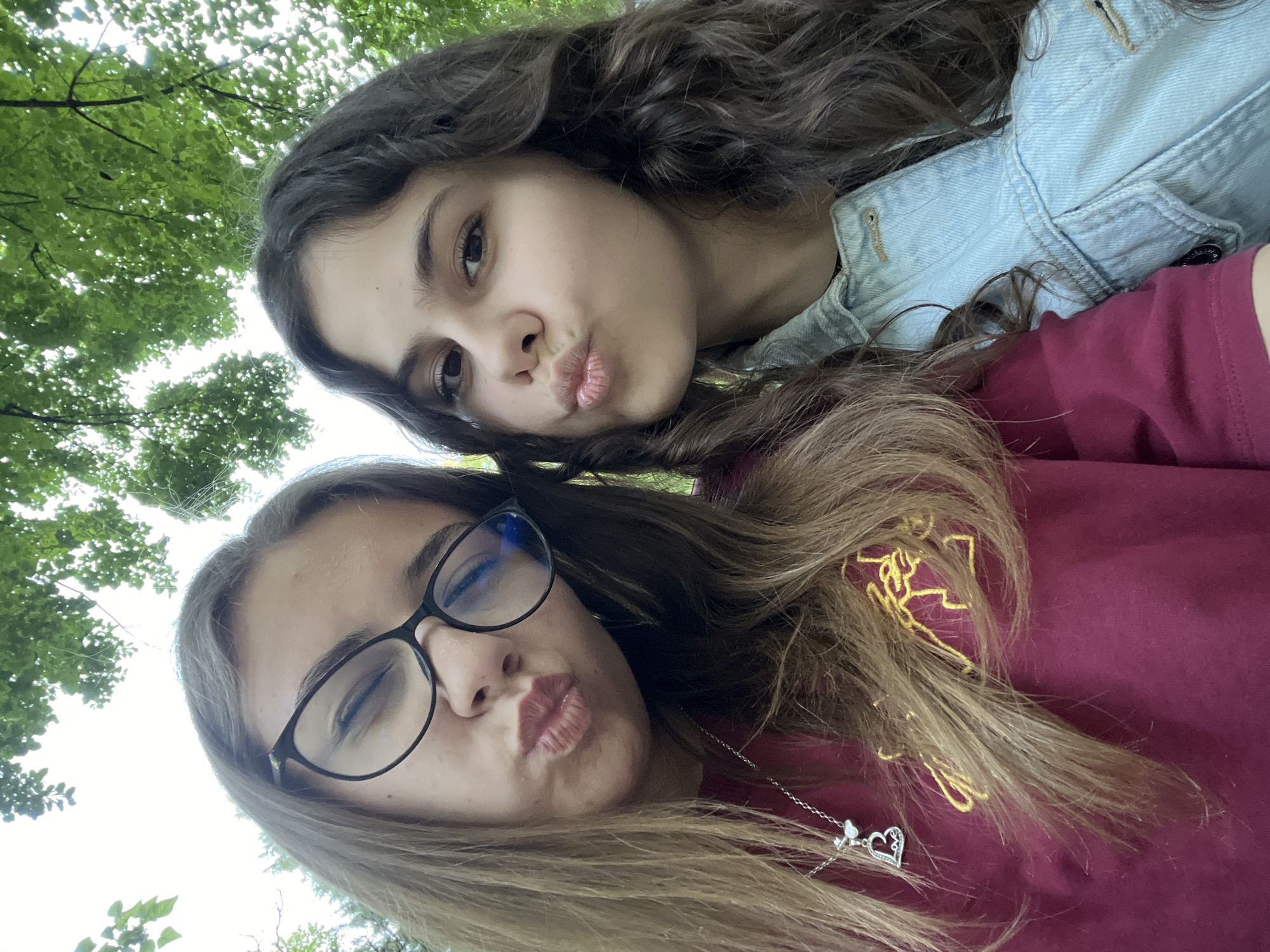 Two young females are posing closely together for a selfie outdoors. The environment shows trees and foliage in the background, indicating a park or green space. The photo was likely taken from a low angle, looking upwards at the subjects. One female, with dark curly hair and a denim jacket, is positioned slightly behind and above the other. The other female, wearing glasses and a maroon shirt with embroidery, is slightly below. Both females are puckering their lips in a "kiss" pose. A heart-shaped pendant on a silver chain is visible on the female in the maroon shirt. There is no way to specifically identify location in Plovdiv, Bulgaria.