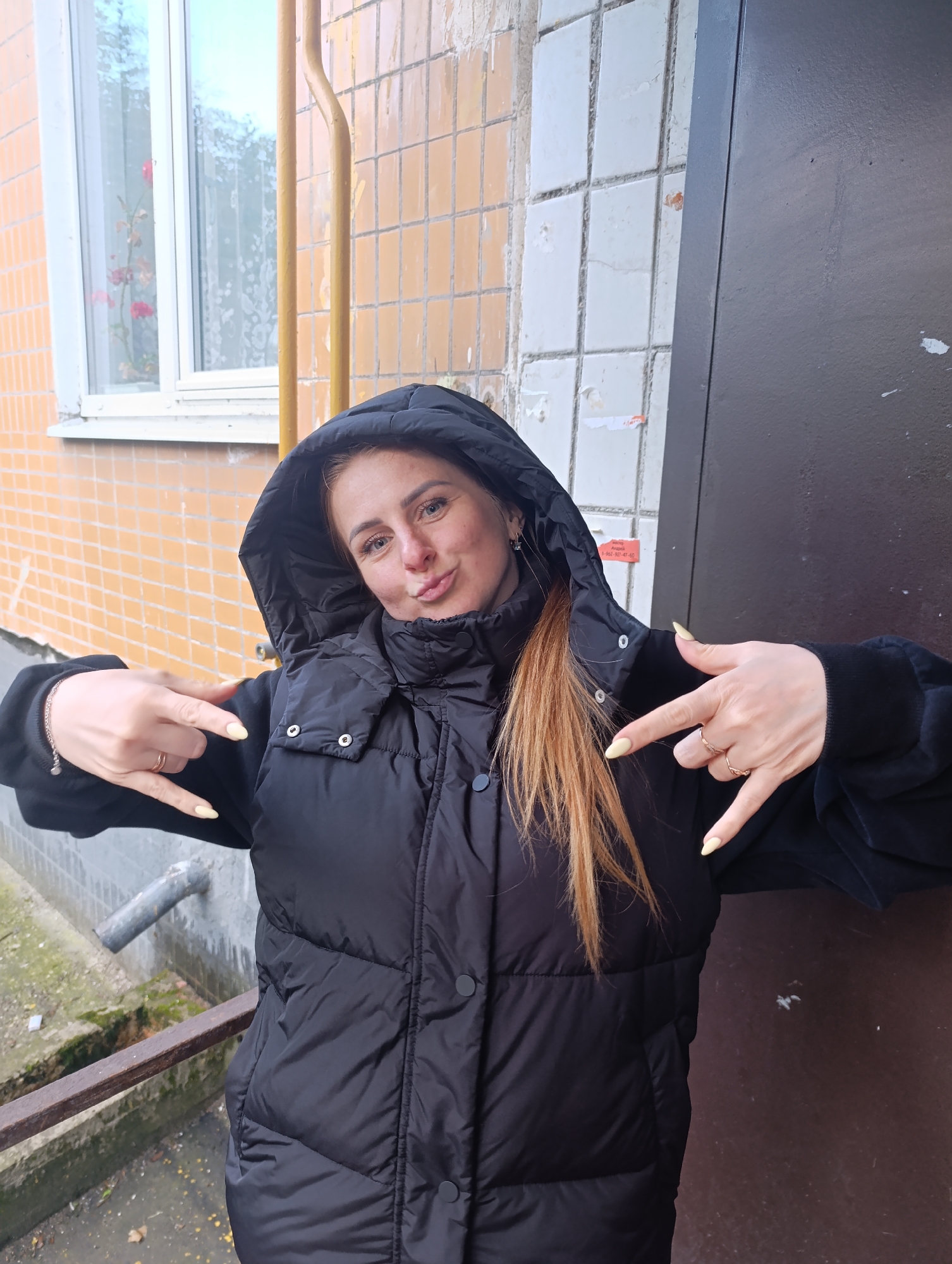 A woman stands in front of a building, posing for a photo. She is wearing a black puffer jacket with the hood up, and her red hair is visible beneath the hood. She is making a "rock on" hand gesture with both hands.
The building behind her has a mix of orange and white tiled walls, a window with flowers in the background, and a dark brown door on the right. A metal railing is visible in the foreground. The location appears to be an urban area.