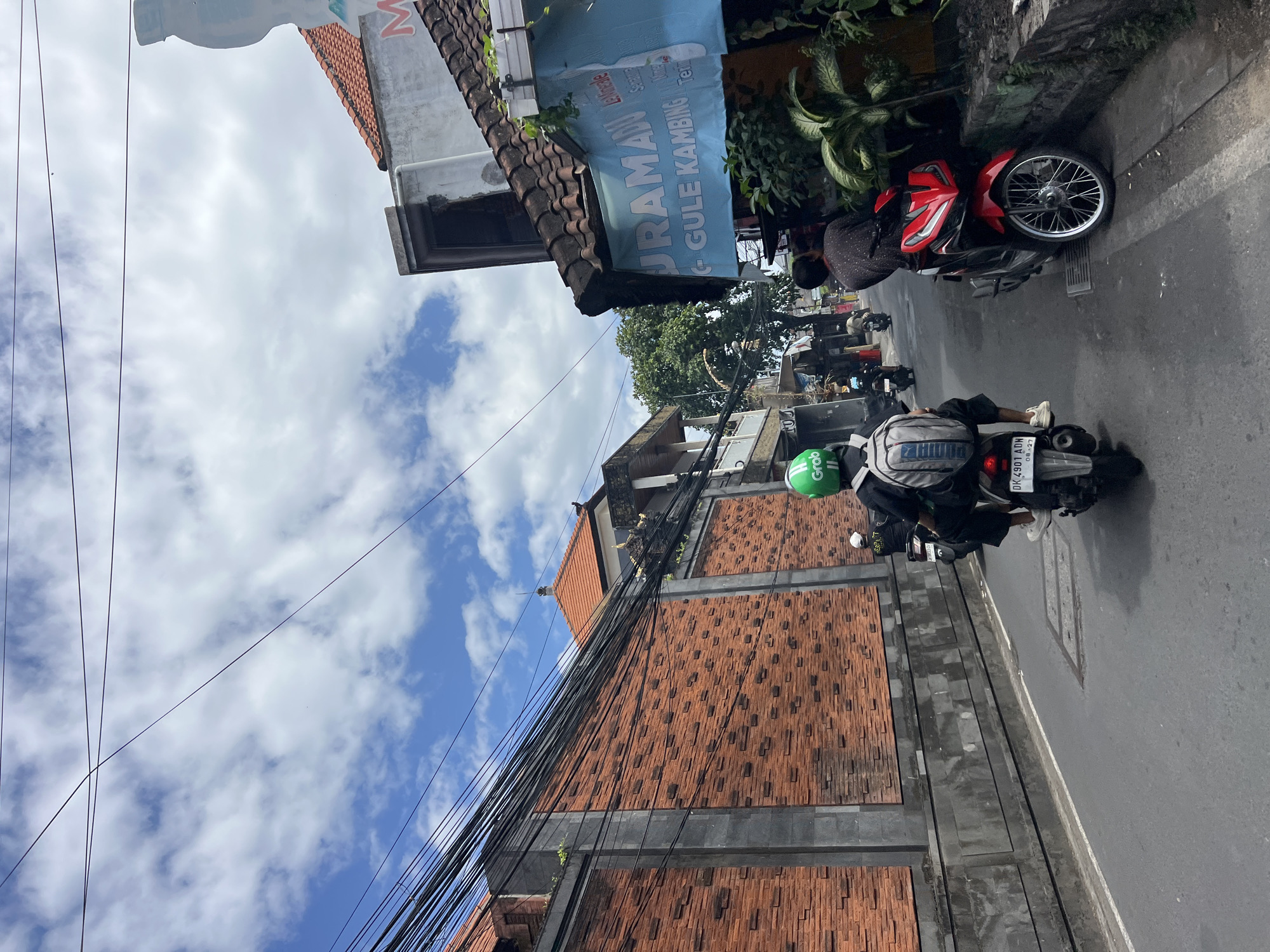 In Kuta, Indonesia, an overcast sky with white clouds is visible above a street scene. A brick wall, approximately 8-10 feet high, runs along the left side of the road, with a grey concrete border at the top. Power lines are strung overhead. On the right side of the street, a blue banner hangs above a shop with red-tiled roofs. The banner reads "DURAMAN GULE KAMBING". Several people are riding motorcycles. In the foreground, a person wearing a green helmet and a backpack rides a motorcycle, with the back of the vehicle visible. Further down the street, another person on a red motorcycle is parked on the right side, facing away from the camera. More motorcycles and people are visible in the distance.
