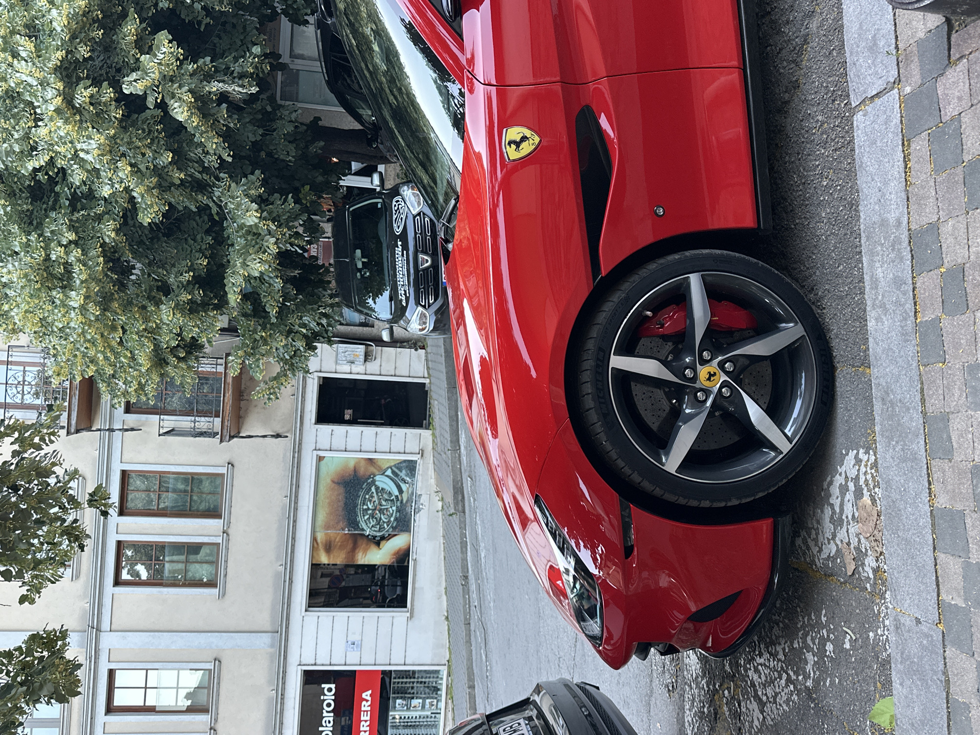 This is a medium shot, eye-level, color photograph taken outdoors. A bright red Ferrari is parked on a cobblestone street, angled towards the viewer. The car's front fender and wheel are prominently featured, showcasing its sleek design and a yellow Ferrari emblem on the side. In the background, a light-colored building with multiple windows and a balcony stands. A large tree with lush green leaves partially obscures the building. Further in the background, a dark SUV is parked, and a portion of another vehicle is visible on the left. The scene appears to be in a city, likely in Veliko Tarnovo, Bulgaria, given the architecture and the parked vehicles. The lighting suggests it is daytime.