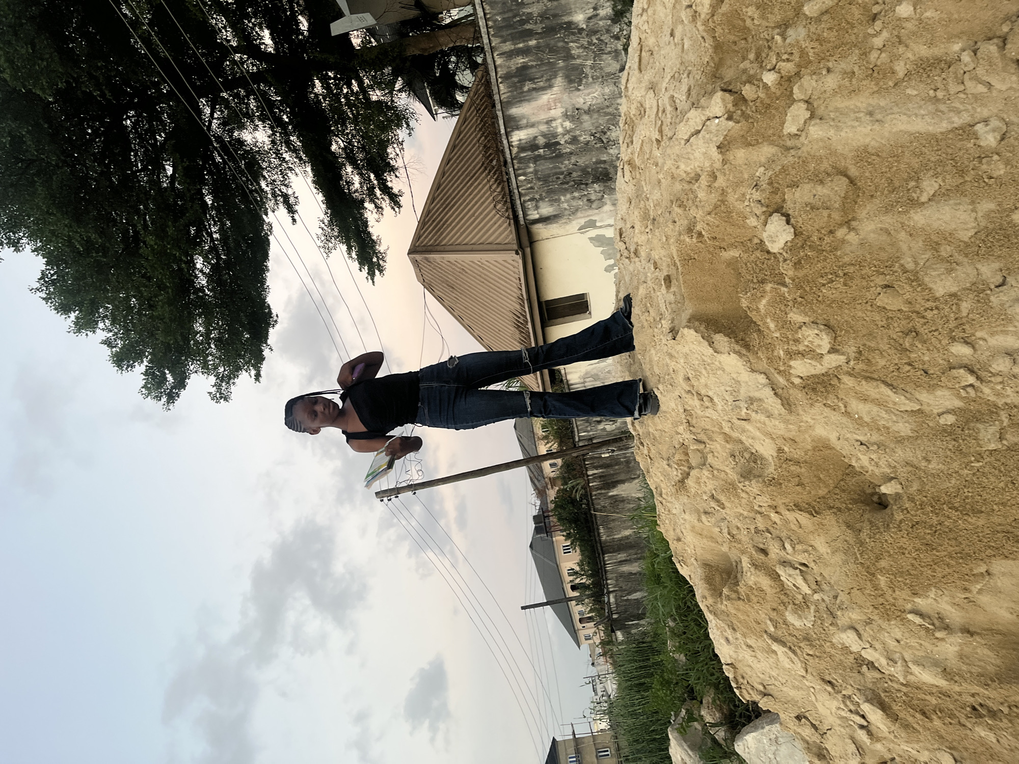 A young individual, appearing female, stands atop a large mound of sand in Iranla, Nigeria. She wears a black sleeveless top, dark blue flared jeans with distressed areas, and dark shoes. Her hair is styled in braids. In her left hand, she holds a small book or notebook with a visible green cover, while her right hand is raised towards her head. The sky above is overcast with light grey and white clouds.
In the mid-ground, multiple utility poles with strung electrical wires traverse horizontally. To the right, a large tree with dense green foliage extends into the upper frame. Behind the sand mound, a concrete perimeter wall, weathered and discolored, is visible, topped with a coil of barbed wire on the right side. Behind this wall, a light-colored, single-story building with a brown corrugated metal roof is present. Further to the left in the background, other residential-style buildings with dark roofs and light-colored walls are visible, one appearing to be a multi-story structure. Green vegetation is interspersed between the sand pile and the wall. The foreground consists entirely of the granular, light-brown sand mound, showing uneven surfaces and small depressions.