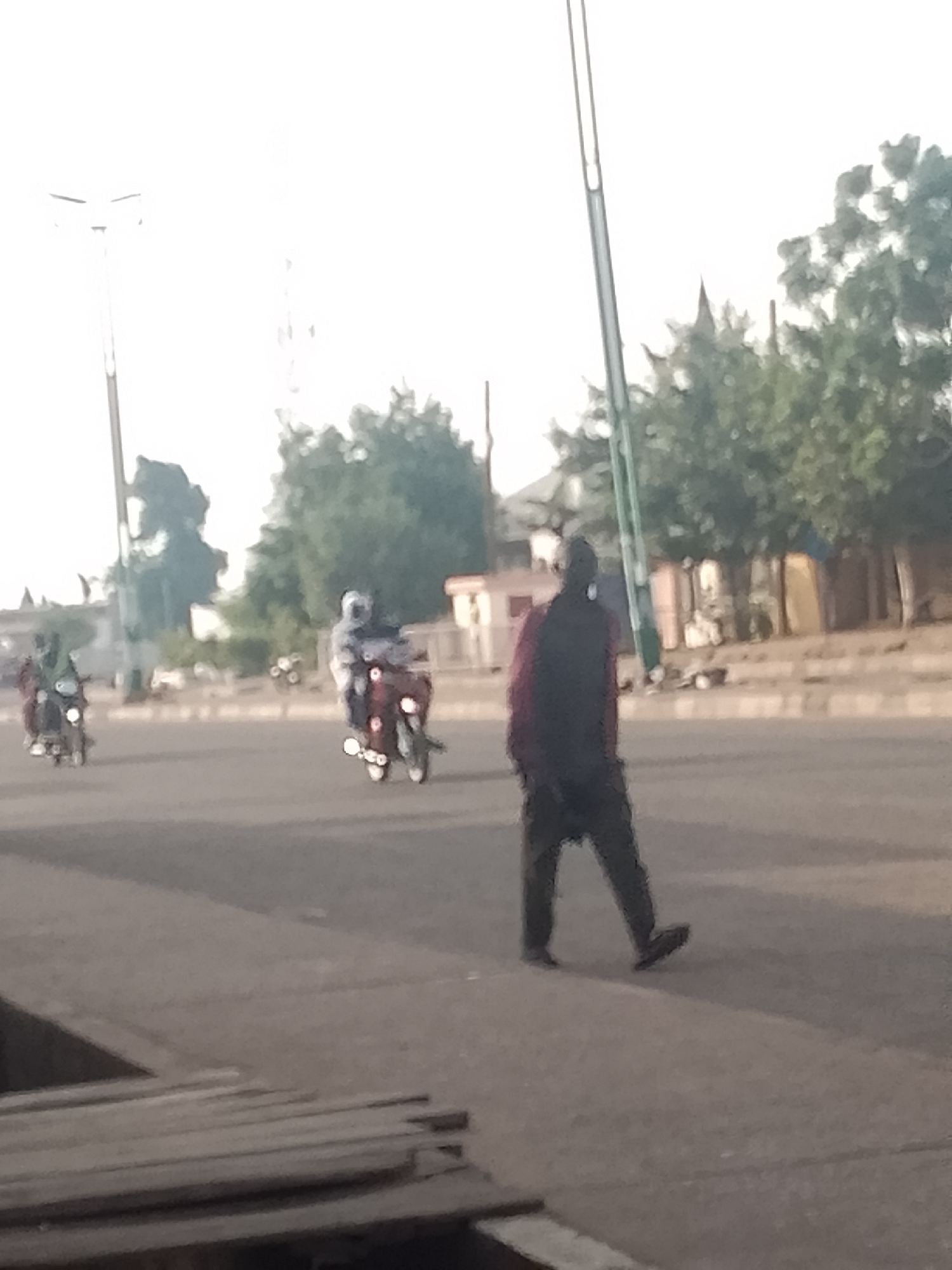 This is a low-resolution, somewhat blurred image of a street scene, likely in Pantame, Nigeria. In the foreground, a person is walking away from the camera, wearing dark pants and a dark jacket with a red scarf or garment visible underneath. The person is on a paved road. Further down the road, two motorcycles are visible. The motorcycle on the left appears to have a rider wearing a helmet. The motorcycle on the right has a rider who is also wearing a helmet and dark clothing. The background shows trees and buildings, some of which are light-colored. A tall, slender pole, possibly a lamppost, is visible on the left side of the frame. The lighting suggests it is daytime, possibly early morning or late afternoon, with the sky appearing bright and washed out. There are some wooden planks or structures in the extreme lower left corner of the image.