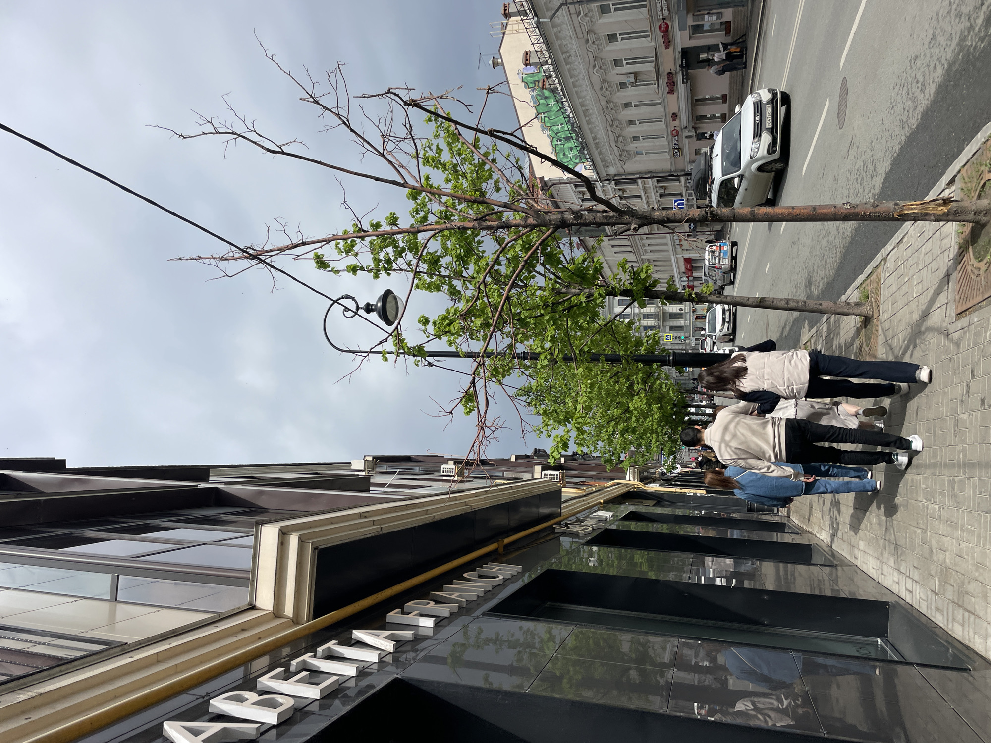 The image depicts a street scene in Kazan, Russia, with a group of three people walking along the sidewalk. They are dressed in casual clothes; one person is wearing a beige jacket and black pants, another is in a light-colored jacket, and the third is in a blue denim jacket and jeans.
The background features a street with vehicular traffic. Buildings are visible, with one identifiable building with the name "TA RANCE" in white lettering on a dark facade. A tree with some green leaves stands near a lamppost on the sidewalk. Above, there is a cloudy sky.