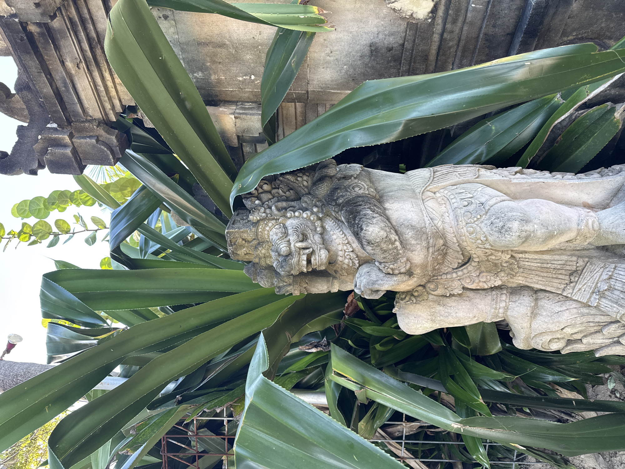 The image shows a stone sculpture of a deity lying amongst green, blade-like plants. The sculpture is heavily weathered. Above the sculpture is a stone structure with carved details. A metal wire fence is visible near the bottom of the image. The location is Kuta Selatan, Indonesia.