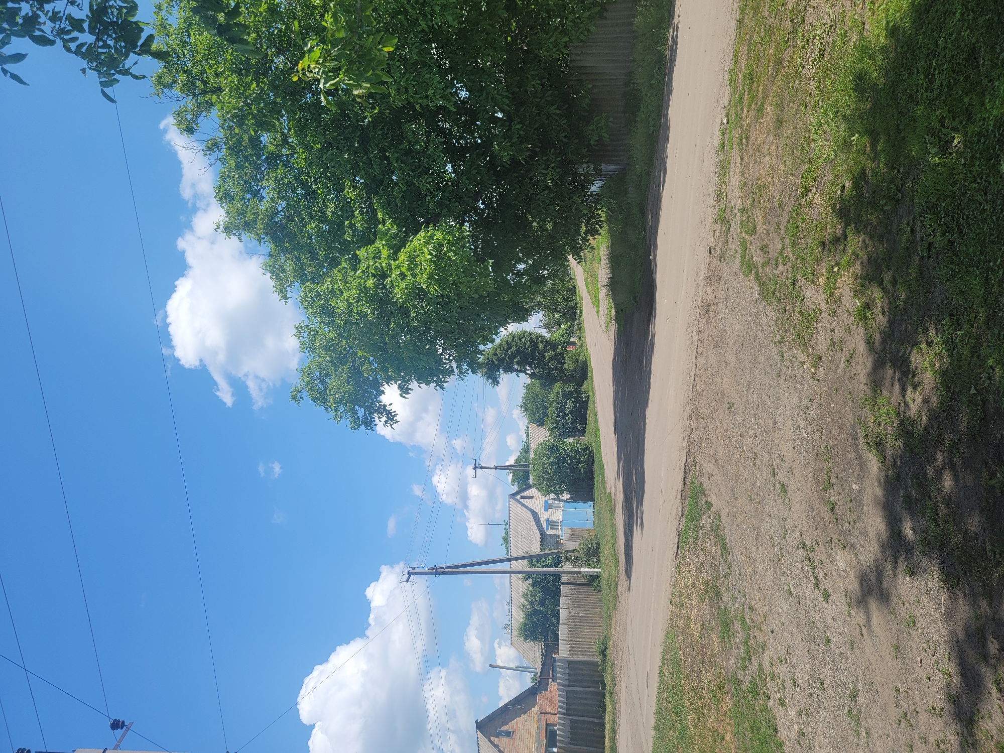The image depicts a rural road intersection on a sunny day in Vovkivka, Ukraine. The road surface is a mix of gravel and dirt, with some patches of grass growing along the edges. In the foreground, the ground is uneven and covered in sparse vegetation and gravel, with shadows from trees cast across it.
To the left of the road, a weathered wooden fence runs along the property line of a residential area. Several houses are visible behind the fence, constructed with brick and concrete block, featuring grey tiled roofs and some with red brick chimneys. One house on the left has a prominent brick facade and a lighter-colored roof. Adjacent to this house is a bright blue gate. Green bushes and trees are planted around the houses.
Several utility poles with electrical wires are present, with one pole prominently positioned near the center of the image. The sky is a vibrant blue, dotted with fluffy white clouds.
To the right of the road, a large, leafy green tree dominates the scene, its branches extending over the road and casting deep shadows. Beyond the tree, a gray corrugated fence lines the property. Further down the road, more greenery and trees are visible, indicating a more wooded area. The overall scene suggests a quiet, pastoral setting in a Ukrainian village. There are no people visible in the image.