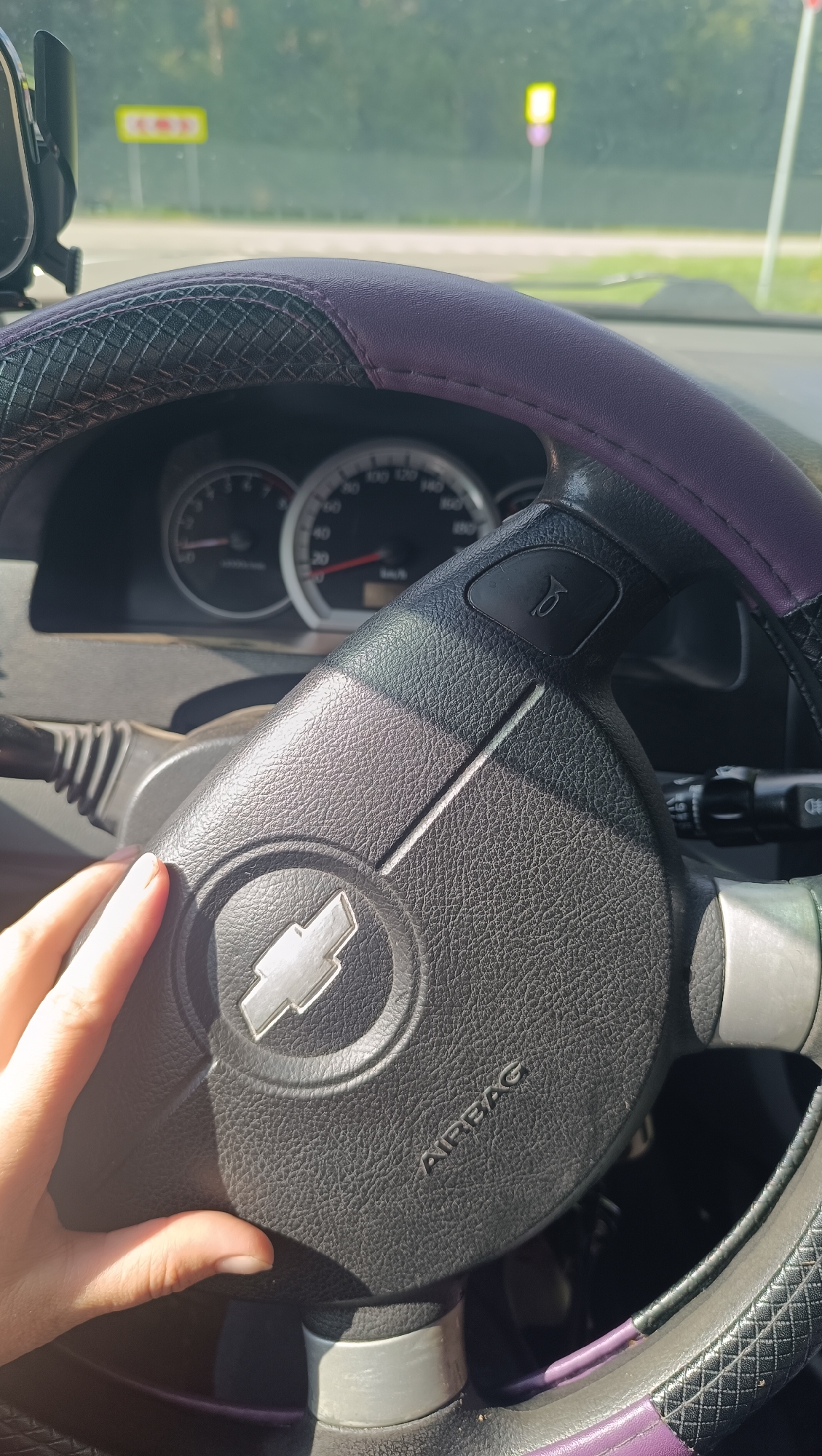 The image shows a close-up view of a car's interior, focusing on the steering wheel. A person's left hand is visible, resting on the steering wheel. The steering wheel is black with a textured grip and a purple trim. A silver Chevrolet logo is prominent in the center of the steering wheel. The word "AIRBAG" is inscribed on the steering wheel. The dashboard and speedometer are partially visible in the background, indicating the car is likely in motion as the needle on the speedometer is above zero. Outside the car, through the windshield, a blurred view of a road and trees is visible, suggesting an outdoor setting. There are also two yellow road signs in the distance, one with an arrow symbol and another with a circular symbol, possibly indicating traffic regulations or directions. The lighting suggests it is daytime. The location is specified as Foki, Russia.