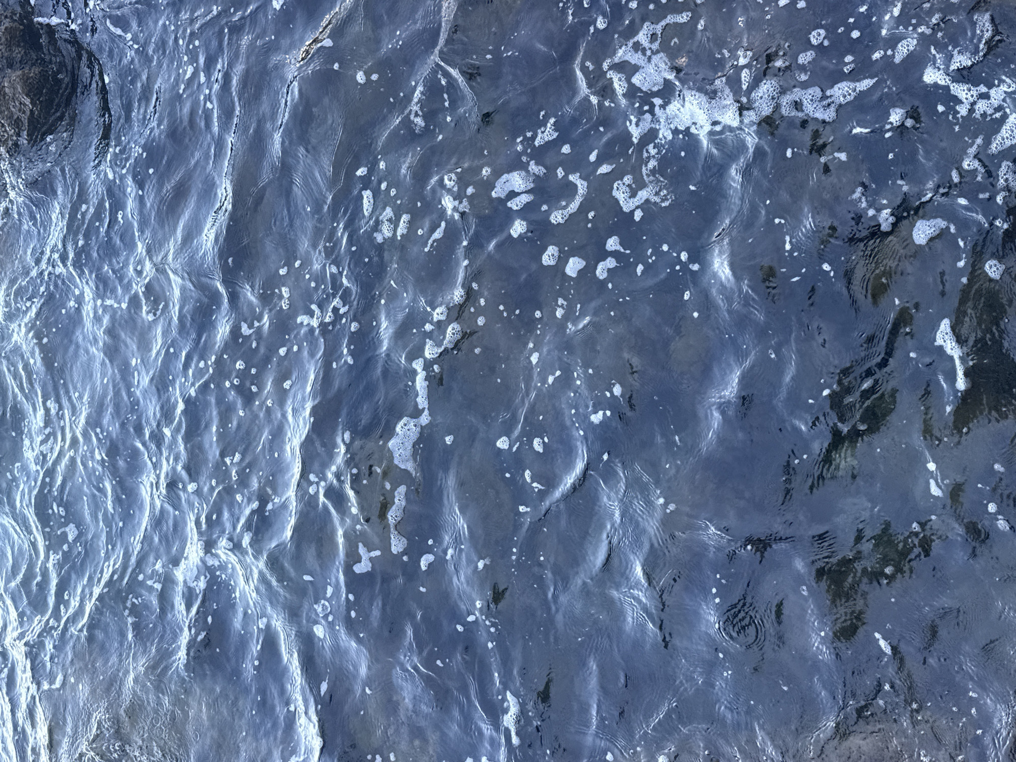 The image shows a close-up, overhead view of dark blue water with gentle ripples and scattered white foam. The water appears to be in a natural setting, possibly a bay or near a rocky shoreline, with a large dark rock visible in the upper right corner of the image. The surface of the water is dynamic, with small waves creating highlights and shadows. Small clusters of bubbles and foam are distributed across the surface, indicating some movement or disturbance. The overall impression is of a calm but active body of water, likely the Adriatic Sea, given the context of Opatija, Croatia. There are no people or specific structures visible in this close-up shot of the water itself. The environment is aquatic, with the water being the primary subject.