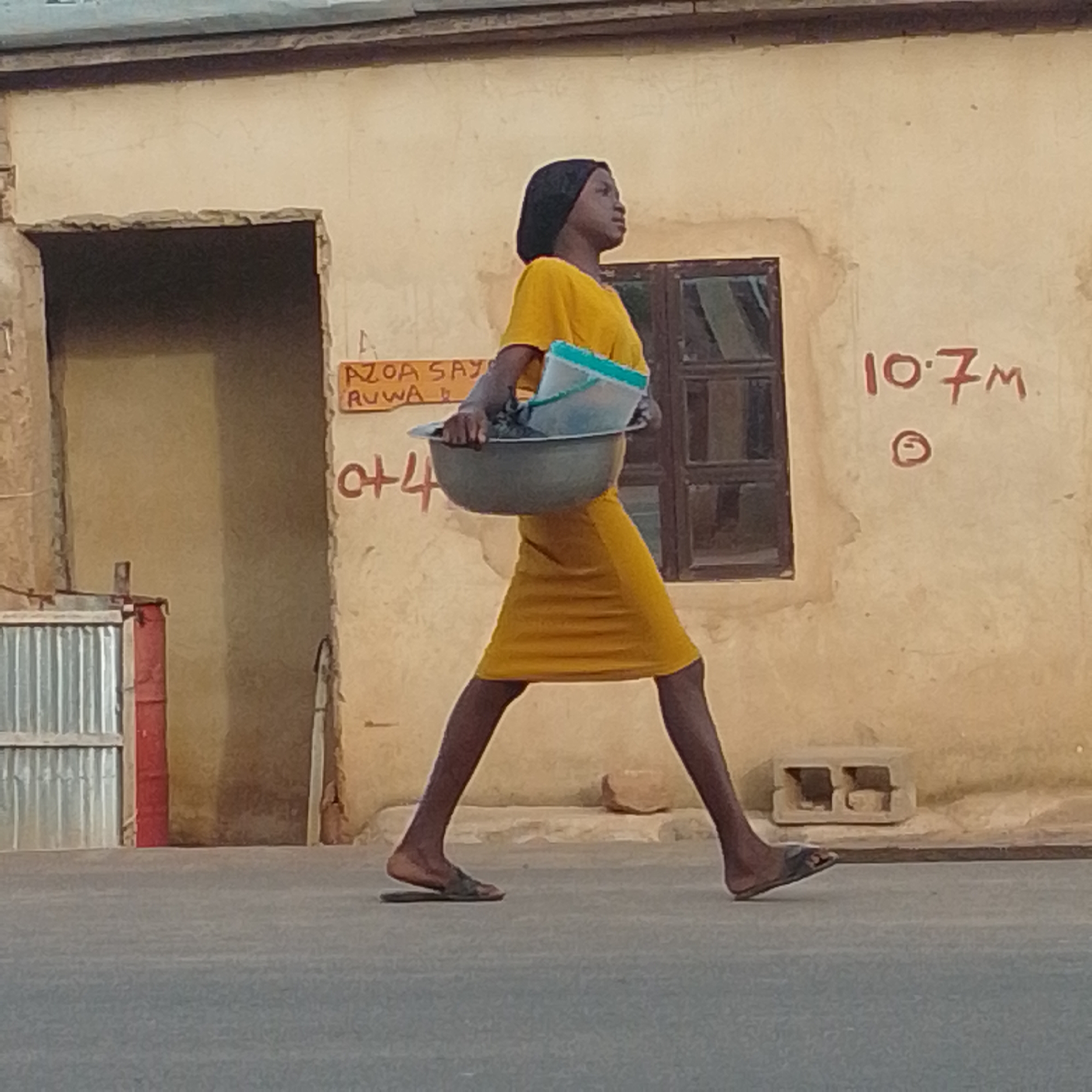 In a street scene in Tempure, Nigeria, a young woman walks in profile from left to right. She is wearing a bright yellow dress and a black headscarf. She carries a large gray basin containing a blue and white plastic container. The background consists of a textured, pale yellow wall with a dark-framed window on the right. Red lettering on the wall includes the text "AZOA SAY RUWA" and "10.7M" with a circular symbol below it. To the left of the woman, a section of corrugated metal fencing and a red cylindrical object are visible. The ground is a gray asphalt surface.
