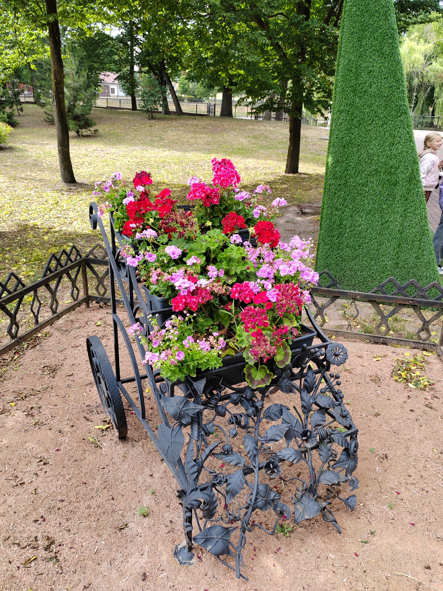 A decorative, wrought-iron, antique-style cart filled with vibrant pink and red geraniums is prominently displayed on a sandy path within a park setting. The cart, with its two large spoked wheels, has an ornate, vine-like design on its base. A low, decorative black metal fence runs along the left side of the cart and extends into the background. Behind the cart, a tall, cone-shaped topiary or artificial green structure stands to the right. The background features a park landscape with trees, a grassy area, and a glimpse of buildings in the distance. A young girl with blonde hair in a braid, wearing a light-colored jacket, stands to the right of the green structure, looking towards the left. The ground around the cart is covered with sand and fallen leaves. This scene is located in Brest, Belarus, likely in a park or garden area designed for public enjoyment.