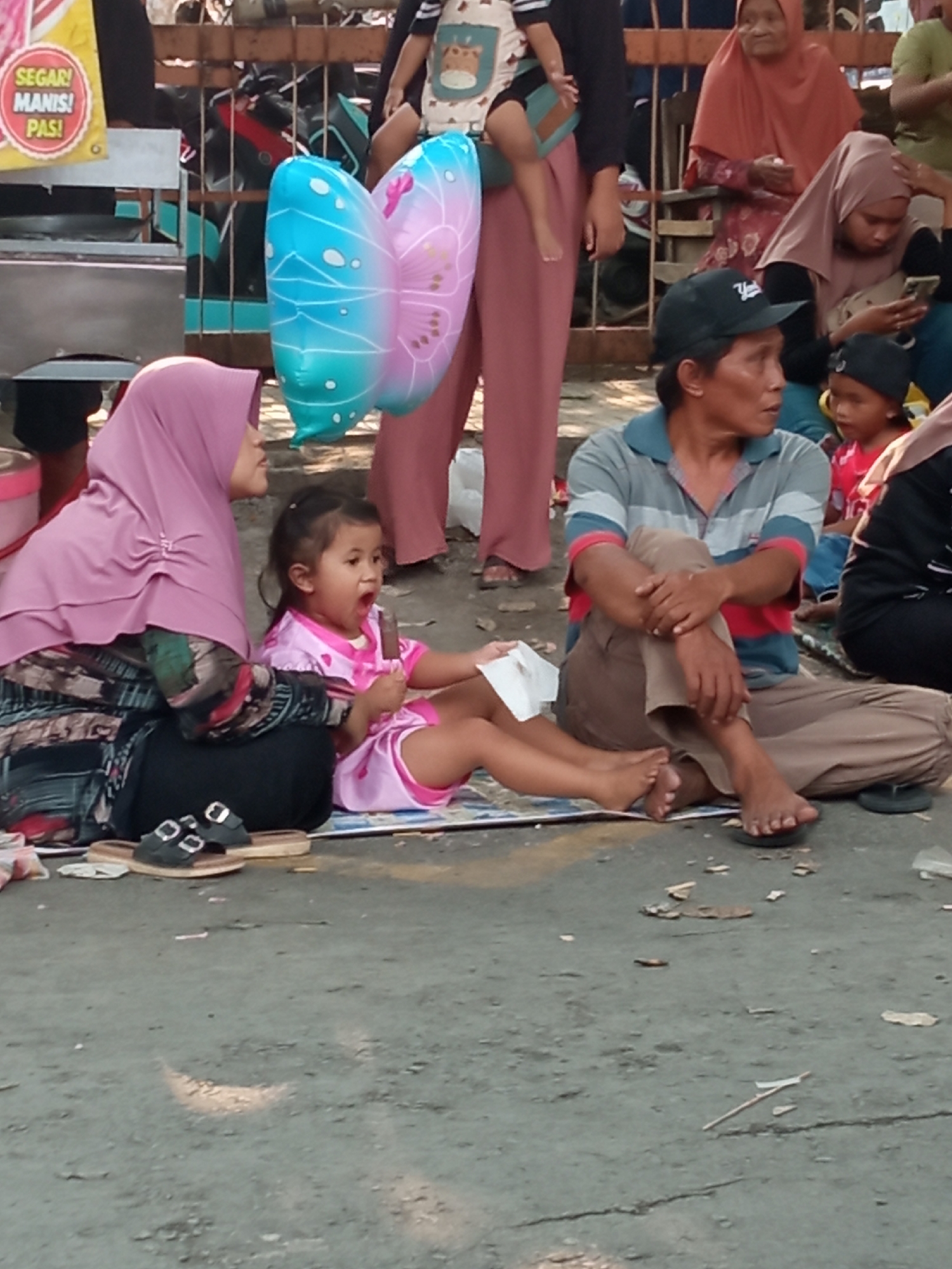 In Balun, Indonesia, a street scene unfolds with a family gathered on the ground. A woman wearing a pink hijab and patterned skirt sits with her legs crossed, looking to the side. Next to her, a young girl in a pink dress with a white trim sits with her mouth open, holding a dark-colored popsicle. In front of them, a man in a grey and red striped shirt and beige pants sits with his legs crossed, his arms resting on his knees. Behind the man, a young boy in a red shirt and a black cap is visible. In the background, other people and various objects, including butterfly-shaped balloons, are present. The ground is a rough, gray surface, likely asphalt or concrete, with some debris scattered around.