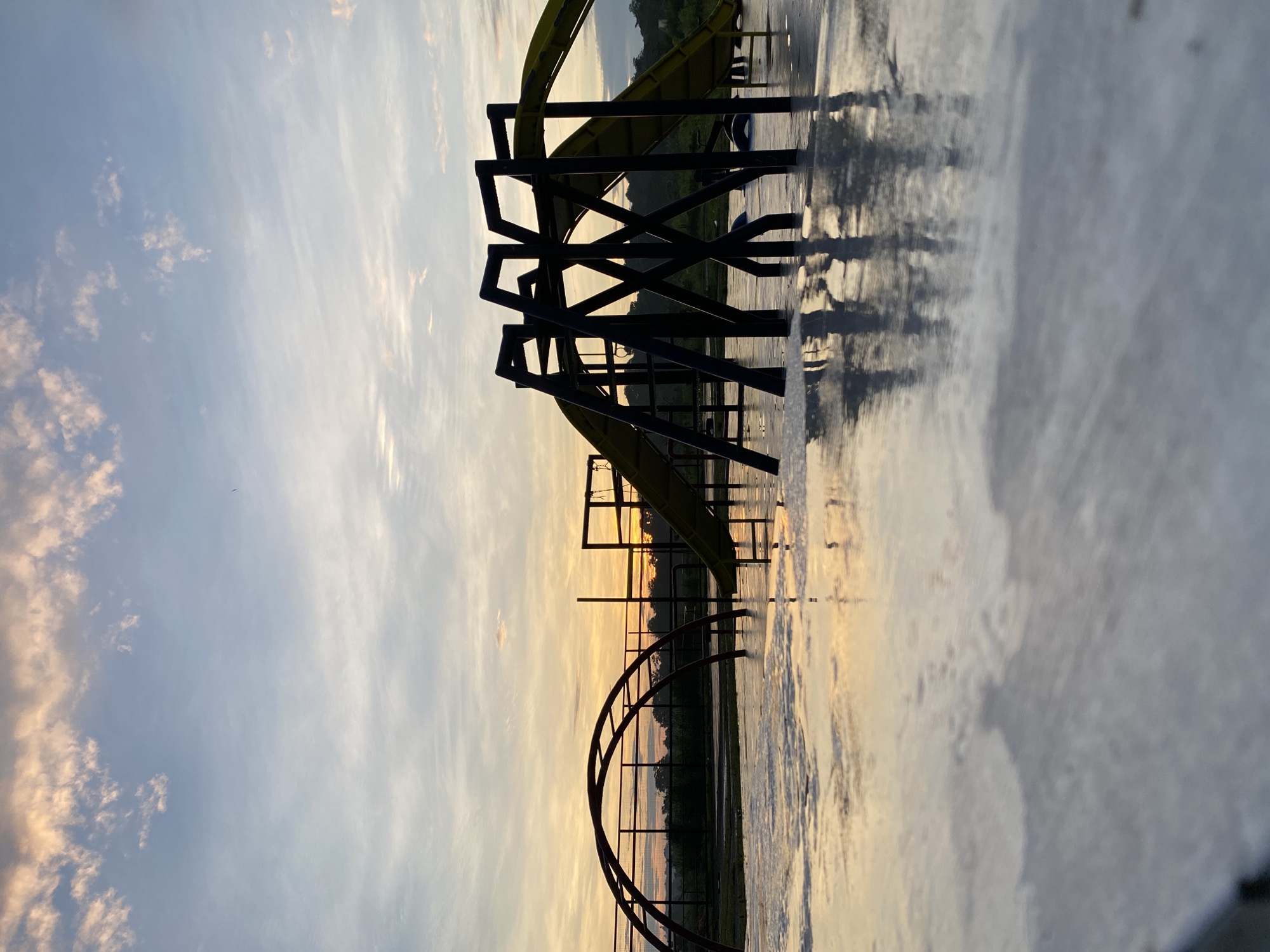 The image, oriented horizontally, depicts a serene outdoor scene at what appears to be a water park or recreational area during either sunrise or sunset. The sky, which occupies the upper portion of the frame, transitions from a soft blue at the top to warm orange and yellow tones near the horizon, indicative of a low sun angle. Scattered, wispy clouds are visible across the sky.
The main subjects are the dark silhouettes of water slides and their supporting structures, standing starkly against the brightly lit sky. In the midground, a large, intricate black metal support structure holds a yellow water slide, which curves through the frame. To the left, another dark, curved structure, possibly a section of a smaller slide or a decorative arch, is also silhouetted.
The ground, which makes up the lower half of the image, is wet and highly reflective, mirroring the sky's warm colors and the dark outlines of the structures. This reflection creates a sense of depth and emphasizes the tranquil atmosphere. Faintly visible in the distant background are the dark outlines of hills or mountains, adding to the scenic backdrop. There are no people visible, suggesting the area is empty, perhaps closed, or captured during a quiet hour. The overall mood is one of stillness and calm. No text is visible in the image. The scene is located in Davao City, Philippines.