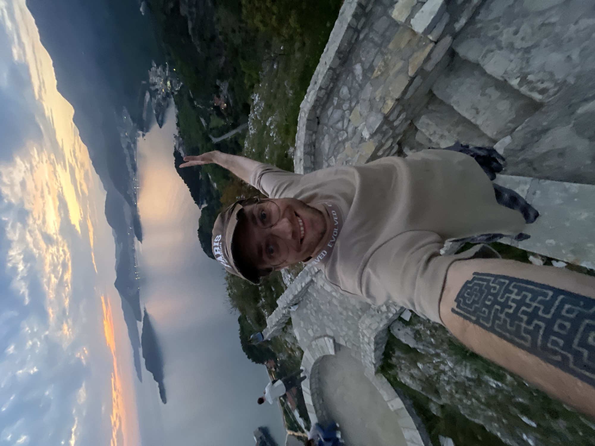 A man is captured in a selfie from a high vantage point overlooking a scenic coastal landscape, likely Sveti Stefan, Montenegro, as indicated by the context. He is smiling broadly, wearing a beige long-sleeved shirt and a baseball cap with "PARIS" visible on it. Text that appears to read "VED EVOLVE" (possibly "EVOLVED" partially reversed) is visible on the collar or neckband of his shirt. A prominent black, geometric, maze-like tattoo covers a significant portion of his left forearm.
The man stands on an old, textured stone staircase or wall, which also functions as a railing, providing a sense of elevation. In the background, a large body of water, presumably the Adriatic Sea, stretches out, reflecting the soft, warm light of either sunrise or sunset. The sky is partly cloudy, displaying orange, pink, and light blue hues. Towering mountains frame the water, and small towns or settlements with twinkling lights are visible along the coast and nestled in the distance, indicating dusk or early morning. A few other individuals can be faintly seen further down the stone structure. The overall scene conveys a feeling of enjoyment and appreciation for the vast, beautiful natural scenery.