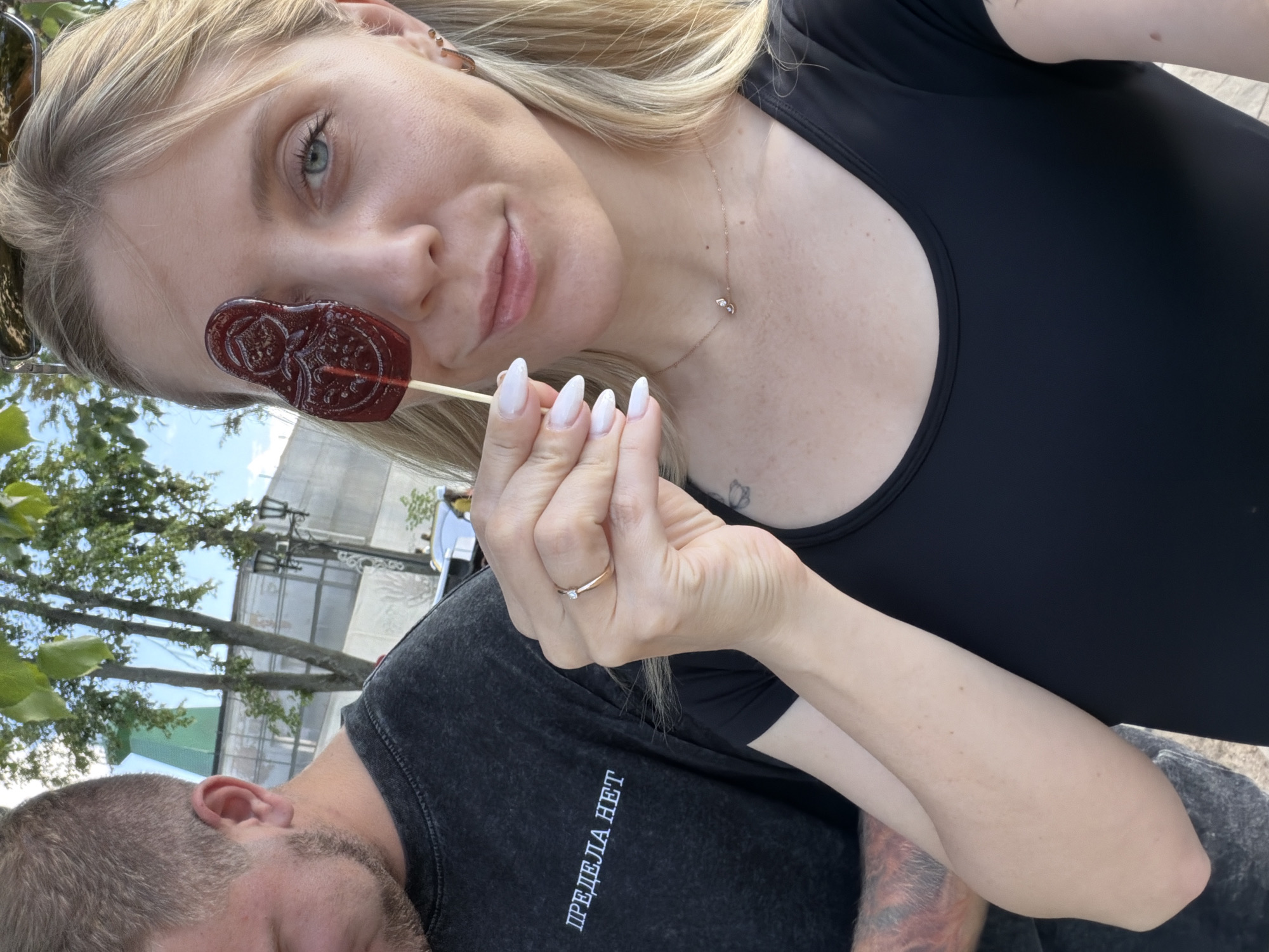 A young blonde woman with fair skin is holding a red, matrioshka-shaped lollipop in front of her right eye, looking at the camera. She is wearing a black t-shirt and a small, delicate necklace. Her nails are painted white. To her left, a man with short brown hair and a goatee is partially visible, wearing a dark t-shirt with white Cyrillic text that reads "ПРЕДЕЛА НЕТ". They appear to be outdoors, with trees and a partially obscured building in the background. The overall atmosphere suggests a casual outing in Suzdal, Russia.