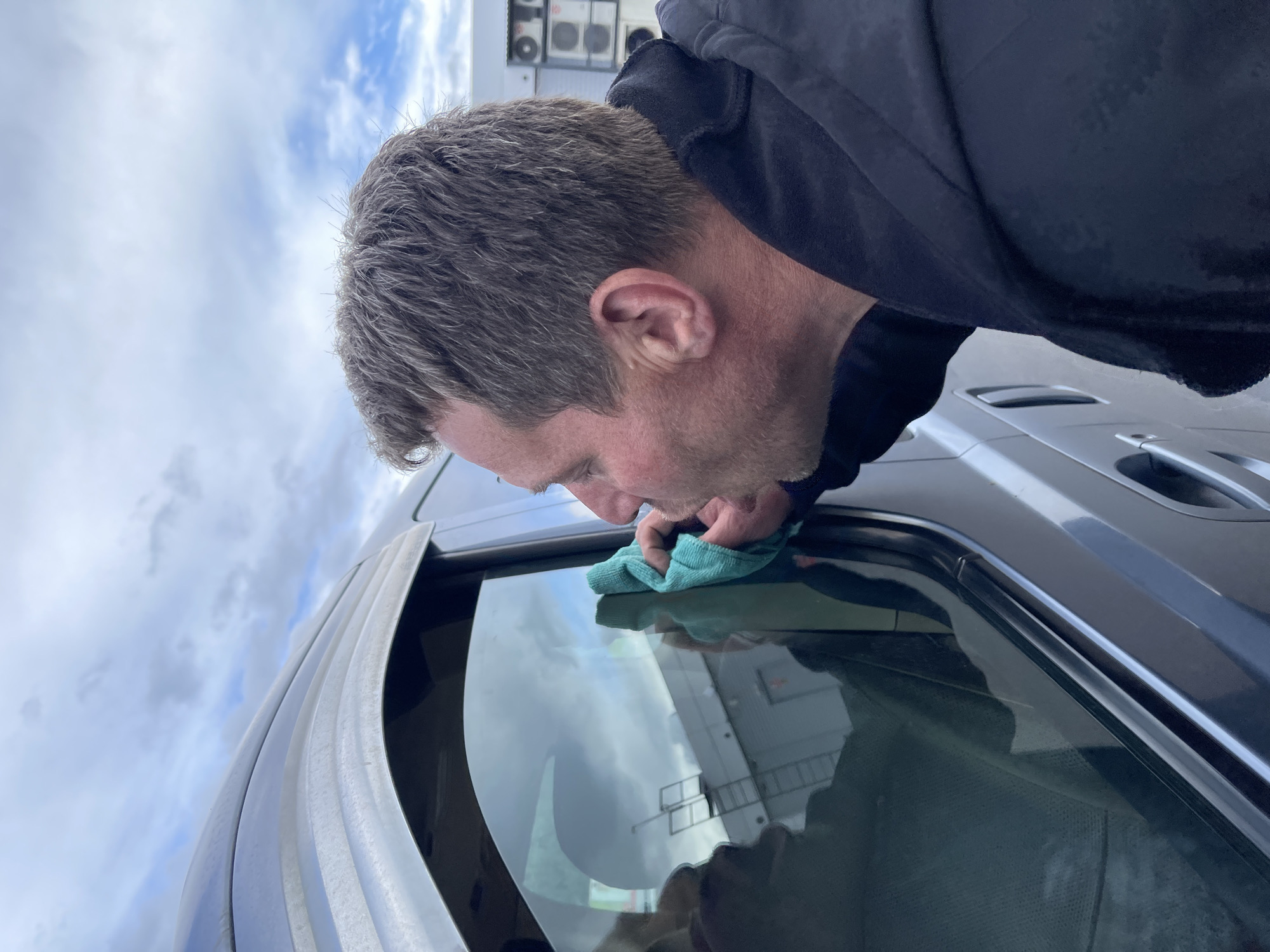 A man is cleaning the window of a dark-colored vehicle. He is holding a blue cloth and appears to be wiping the glass. The reflection in the window shows a building. The sky is cloudy. The location is Бавыкино, Russia.