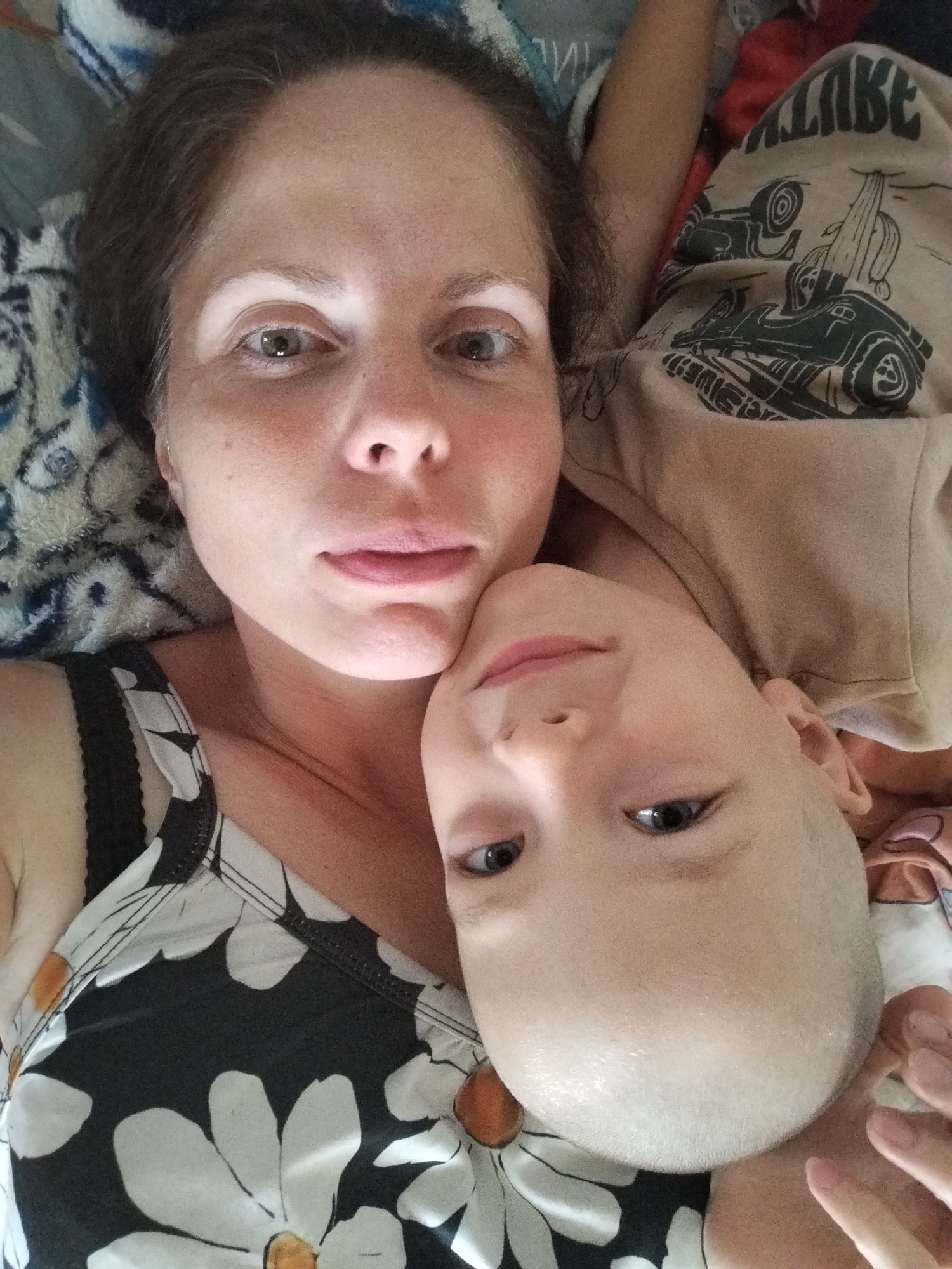 This is a close-up, overhead selfie of a woman and a child lying down. The woman is on the left, her head is tilted back, and she is looking into the camera. She has dark brown hair, fair skin with some freckles, and is wearing a black and white floral tank top. The child is on the right, with their head tilted forward, and is also looking into the camera. The child has fair skin, short blonde hair, and is wearing a light brown t-shirt with a graphic print. They are positioned so that their faces are close together. The background consists of a patterned blanket and bedding. This image was taken in Zavodoukovsk, Russia.