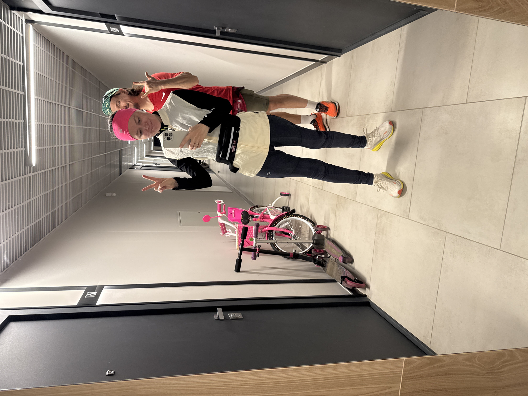 Two individuals stand in an indoor hallway. The first individual wears a pink headband, black long-sleeved shirt, beige jacket, black leggings, and white running shoes with yellow soles. They hold a phone and make a peace sign. The second individual, behind them, wears a green and black baseball cap, red shirt, and green shorts, also making a hand gesture. A pink bicycle and scooter rest against the wall beside the first individual. The hallway has grey doors, tiled floors, and fluorescent lighting. The image was likely taken in Verkhnii Liuk, Russia.