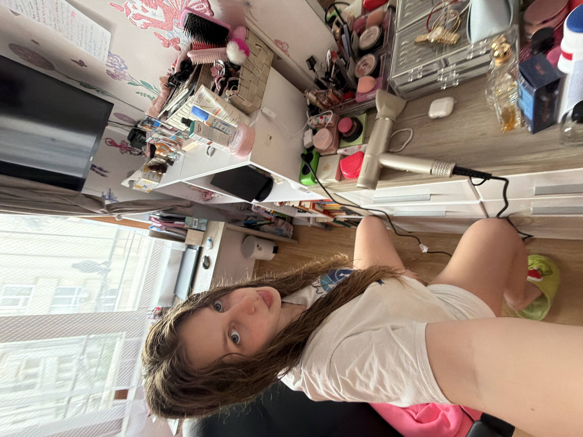 A woman with long, wavy brown hair is seated in a black chair, wearing a white t-shirt and pink bottoms. She is positioned in front of a window overlooking a city. Her legs are visible, and she is wearing a green sandal on one foot. A dark TV is visible on the wall behind her, and the wallpaper has a floral design. There is a white desk crowded with cosmetics, toiletries, a hair styling tool, and other personal items. Based on the architecture visible through the window, this location may be in Burgas, Bulgaria.