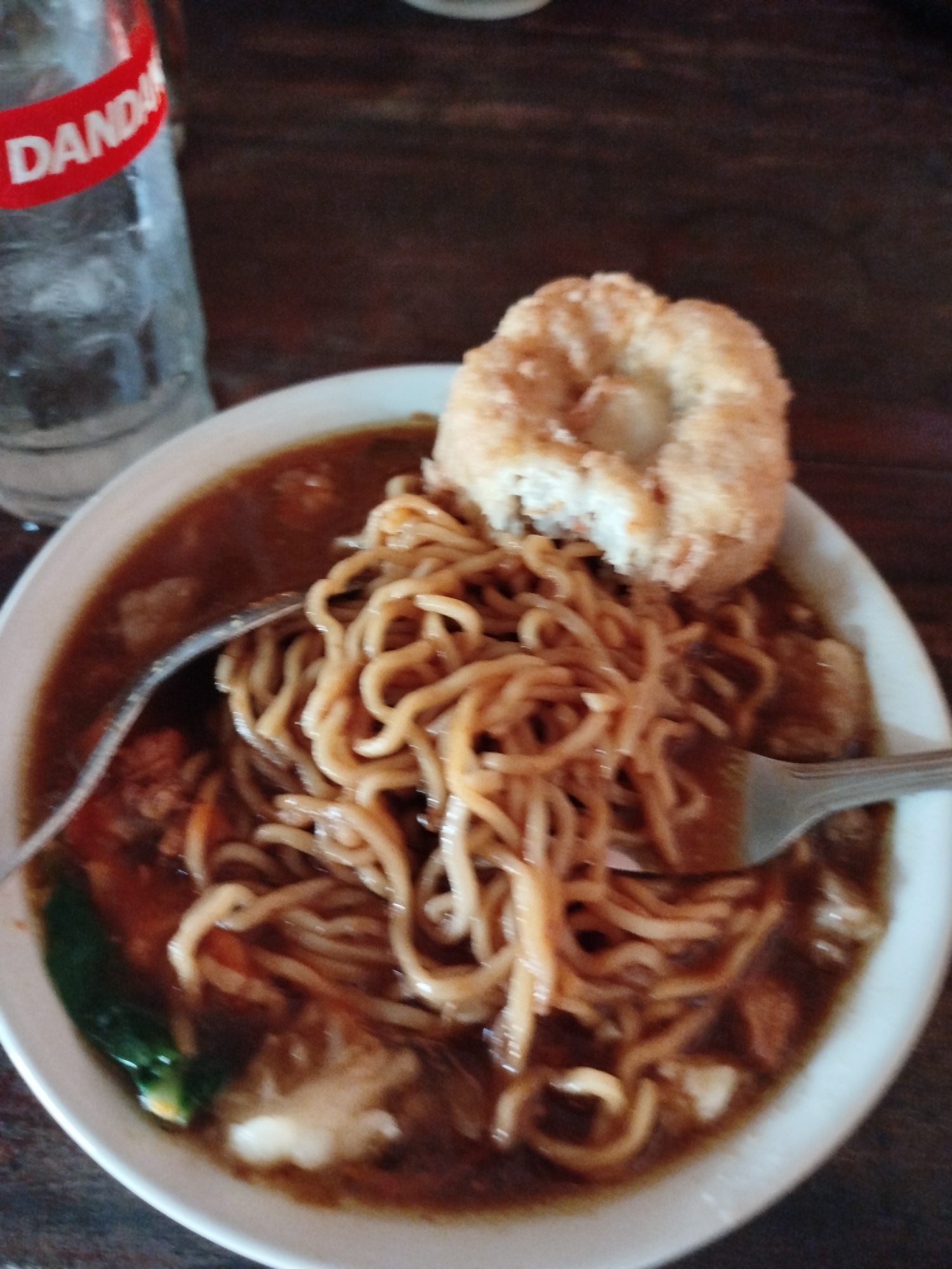 A close-up, eye-level, medium shot captures a white bowl of ramen noodles in a dark broth, topped with a piece of deep-fried food, likely a fritter, in Pojok, Indonesia. A fork rests on the right side of the bowl, partially submerged in the broth. To the left of the bowl, a clear glass with a red label reading "DANDAN" contains ice and a clear beverage. The table surface is made of dark wood. The background is blurred, with only hints of the environment visible.