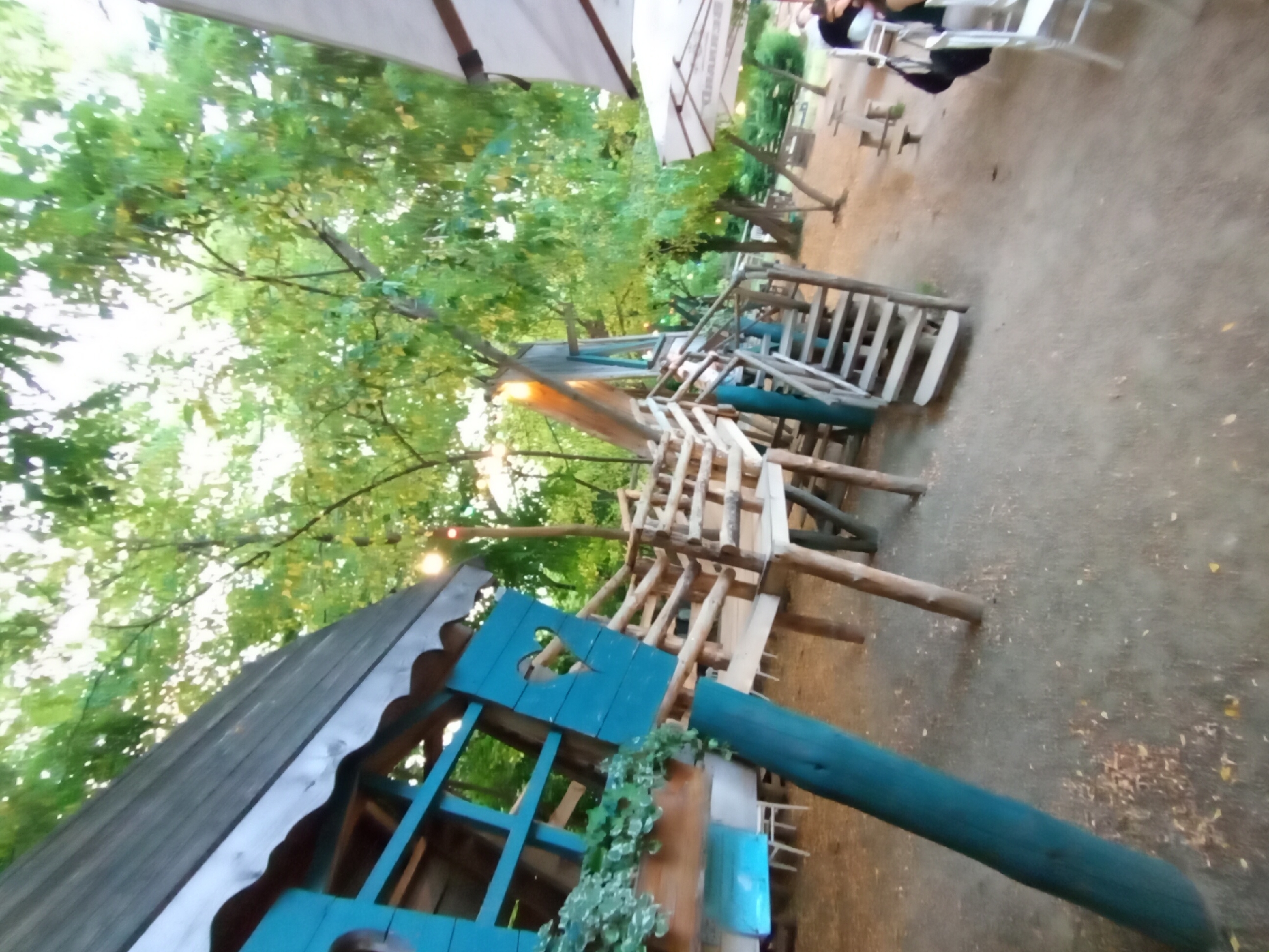 The image captures an outdoor scene, likely a backyard or park area, featuring a rustic wooden play structure. The structure is multi-level, with ramps and ladders made from rough-hewn logs and planks. Some parts of the structure are painted a vibrant teal color, including a small house-like section with a heart-shaped cutout. Green ivy or other foliage is seen growing on and around the structure, adding to its natural, overgrown aesthetic.
In the background, tall trees with lush green foliage create a canopy overhead, suggesting it is daytime. Sunlight filters through the leaves, creating dappled light on the ground. White umbrellas, possibly for an outdoor seating area, are visible in the upper portion of the image. A few scattered wooden tables and chairs are also present, hinting at a space designed for relaxation or recreation.
There are glimpses of people in the background, seated or standing near the umbrellas and tables, but they are not the focus of the image. The overall mood is relaxed and natural, emphasizing the play structure as the main subject within its outdoor environment. The ground appears to be covered in dirt or gravel, with some scattered leaves. The lighting suggests it could be late afternoon or early evening, given the warm tones and filtering sunlight. The scene is likely in a location with a generally mild climate to allow for outdoor activities. No visible text is present in the image.
