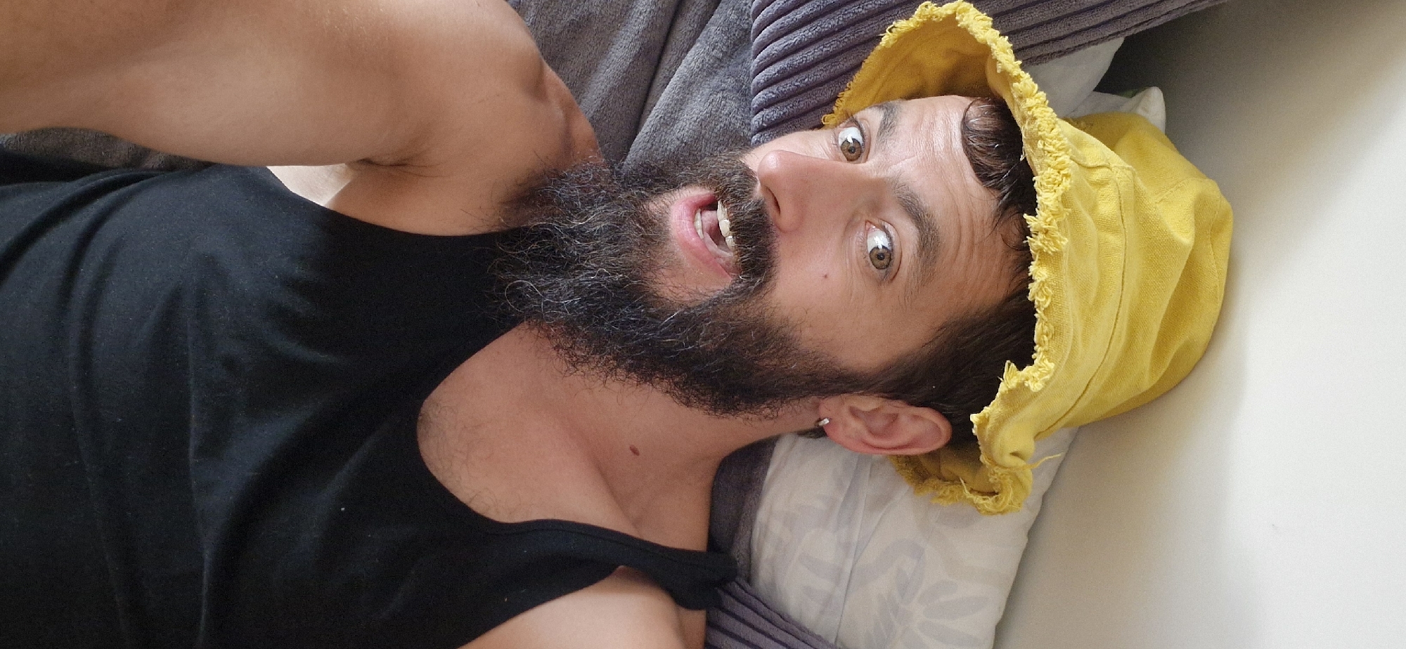 The media file shows a man lying down and looking up at the camera. He is wearing a black tank top and a yellow bucket hat that is tilted over his head. He has a full beard and appears to be smiling with his mouth open. His eyes are wide, and he has a surprised or playful expression. The background consists of pillows and a textured surface, possibly a couch or bed, suggesting an indoor setting. The lighting indicates it is daytime. There is no visible text in the image. The overall mood is casual and humorous.