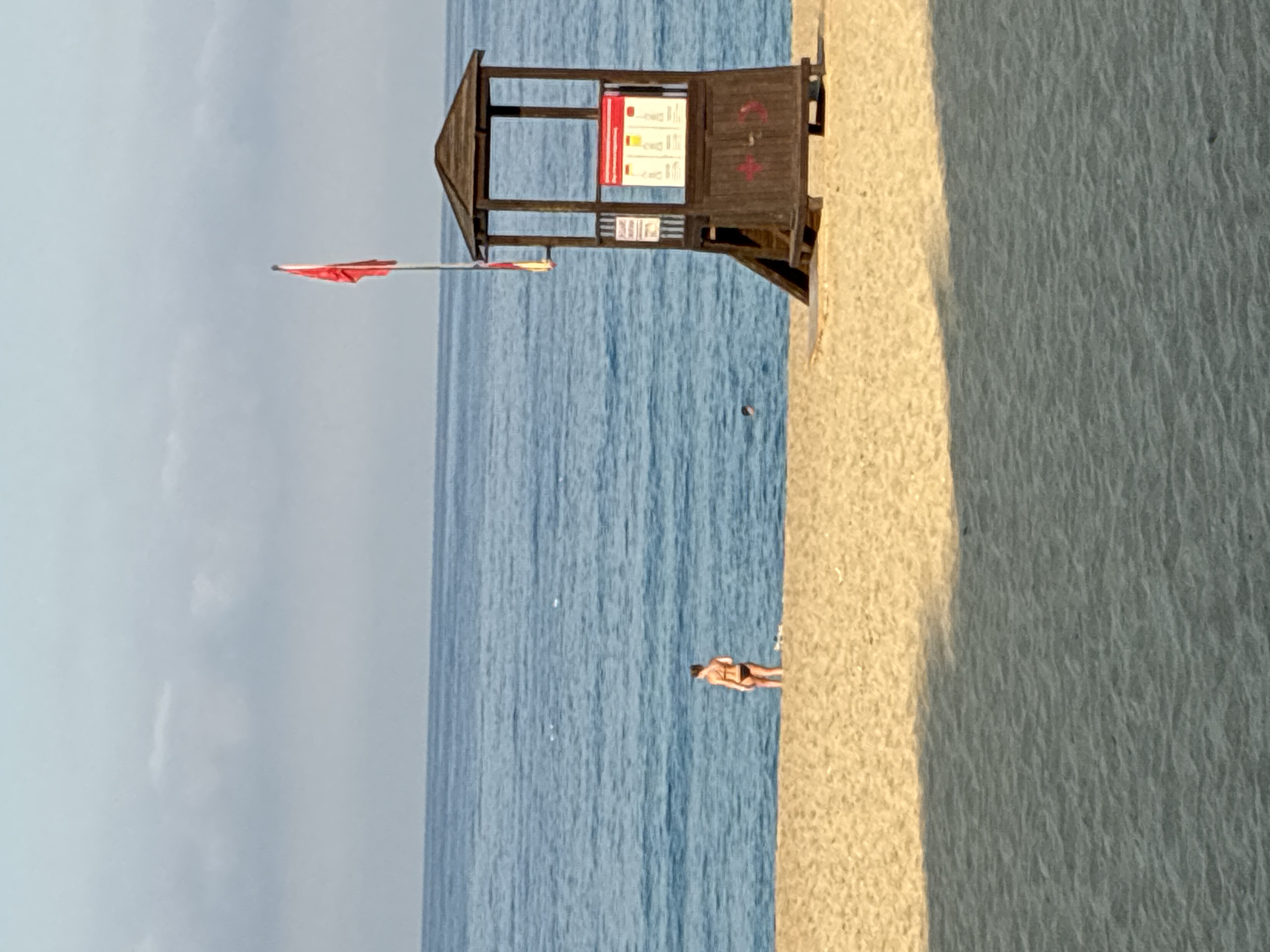 A lone person in a black bikini stands on a sandy beach near the water. To their right, a wooden lifeguard tower stands prominently, flying a red flag. The sea is a calm blue, meeting the horizon under a hazy sky. The beach in the foreground is a light tan, with long shadows cast from an unseen light source.