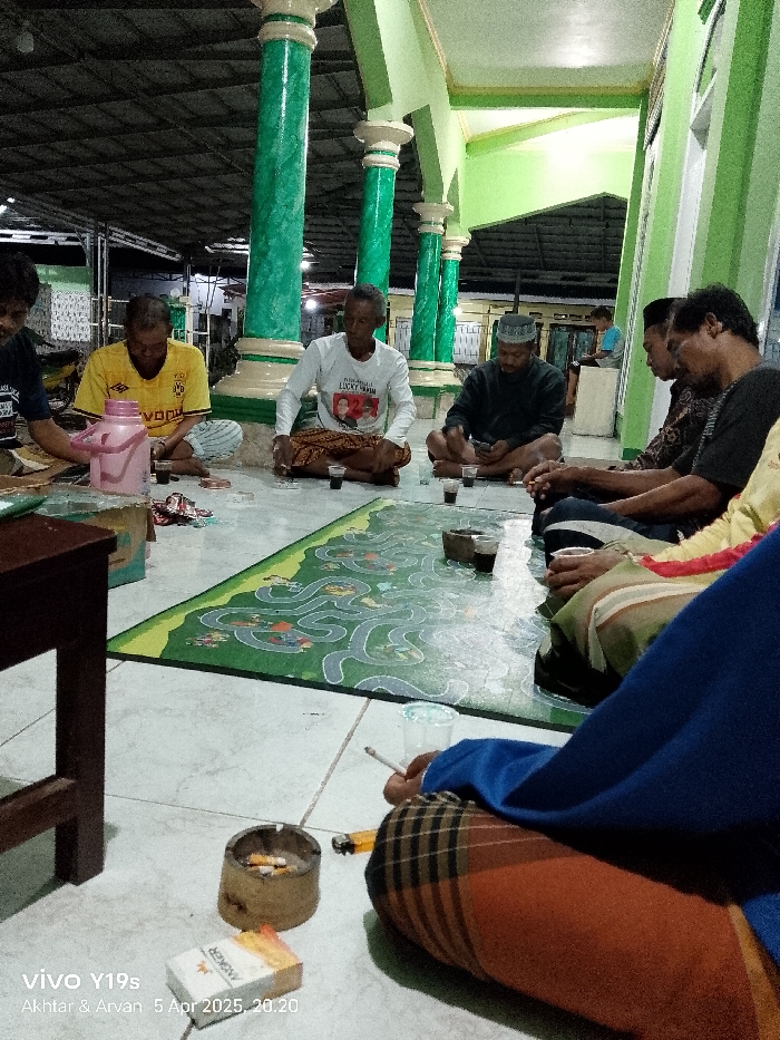 A group of men are sitting on a tiled floor around a green mat with a maze design on it. The men are all wearing casual clothing and some are drinking from glasses. One man is holding a cigarette. They are all sitting in a covered area, which appears to be part of a building with green columns. There is a table to the left of the image. The image was taken in Anjatan, Indonesia, at the coordinates -6.419, 107.979.