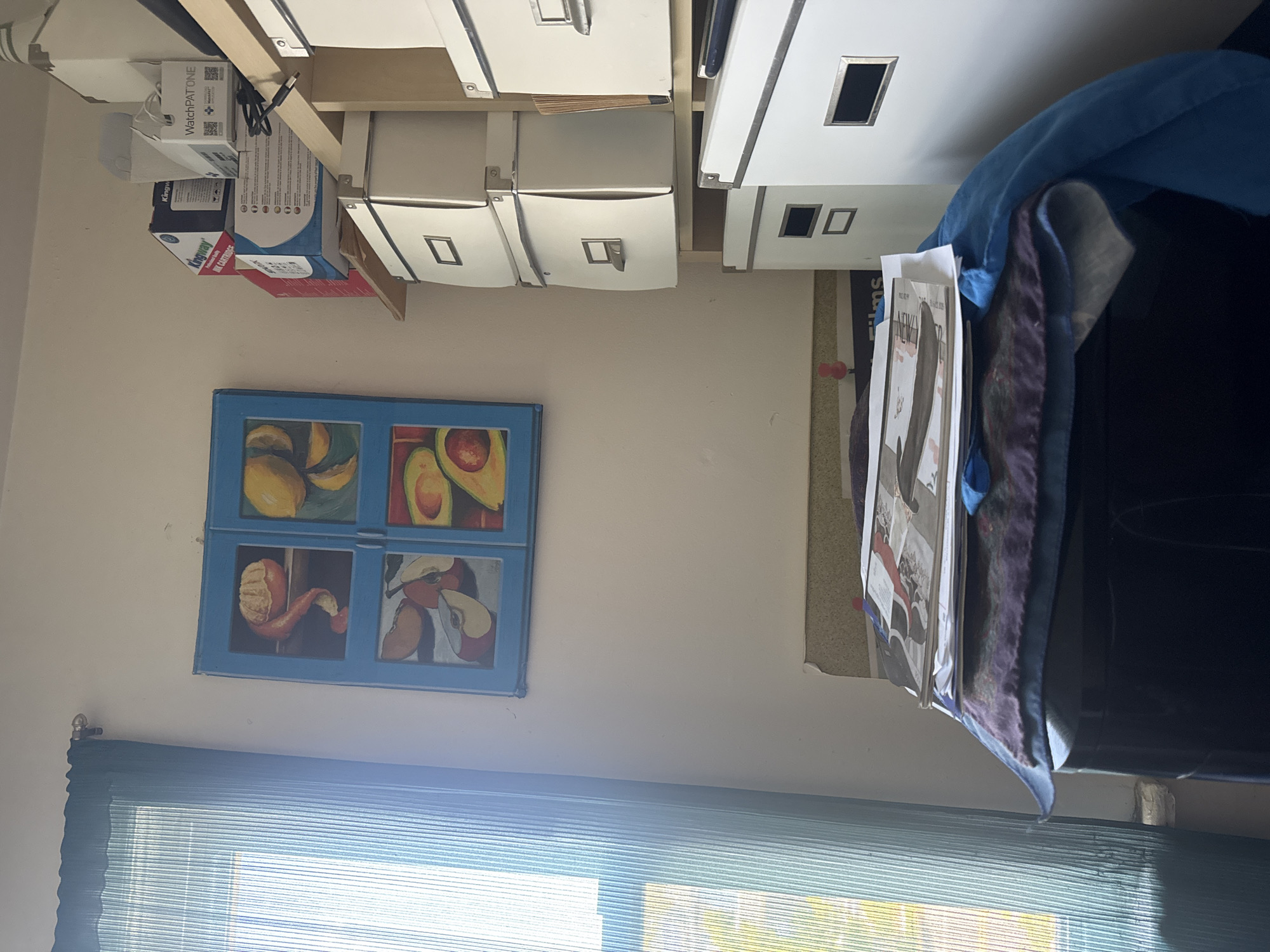 A slightly rotated and angled shot of a room in Neptune Township, United States of America. A white wall is visible with a framed picture showing various fruits on the left. Under the painting, blue curtains are present. A shelf unit runs along the top of the image, and on the right side, there is what appears to be a chair with documents on it.