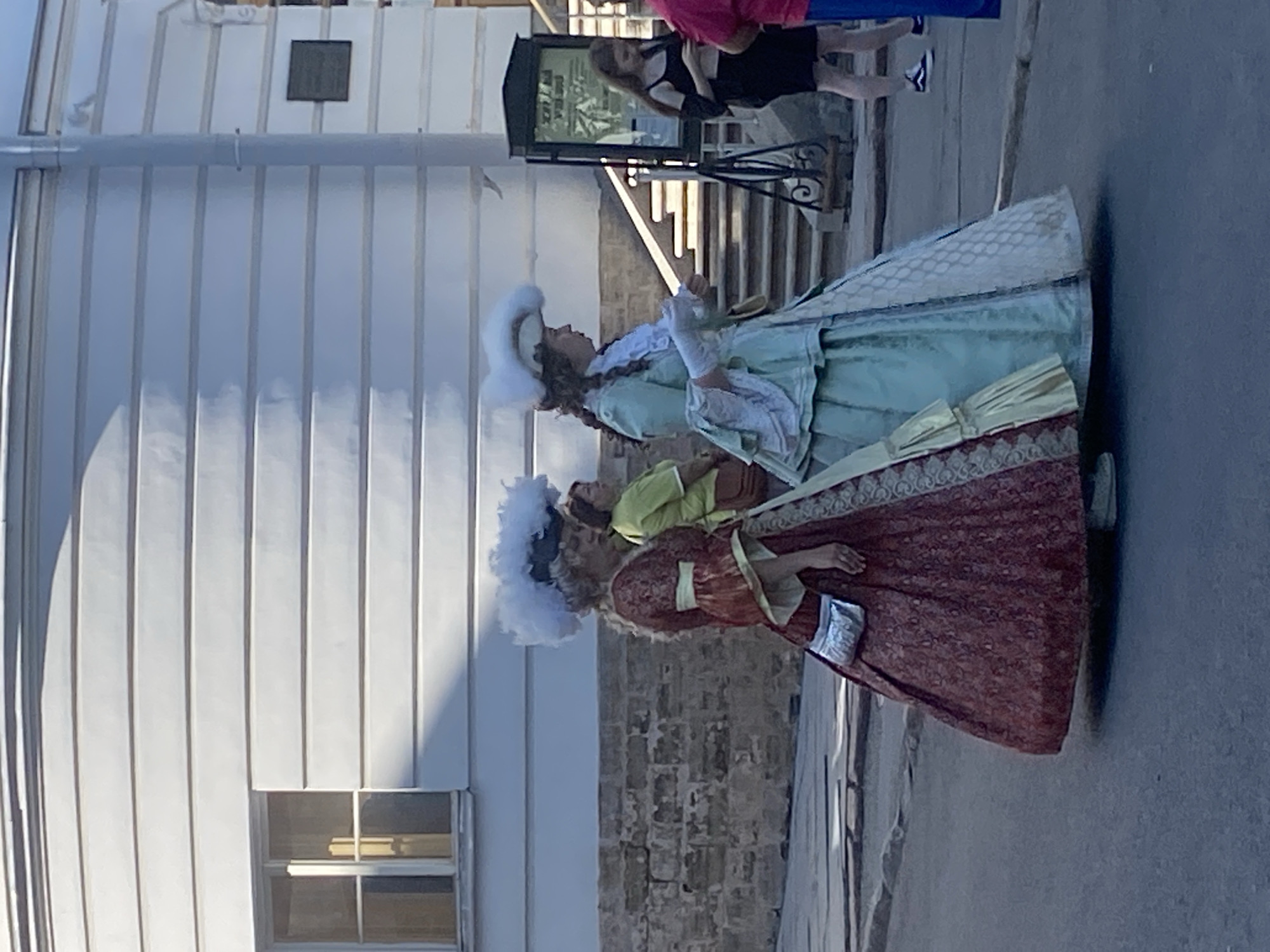 The image shows two women dressed in historical costumes, possibly reenactors or performers, standing on a paved area in front of a white building with horizontal siding. The woman on the left wears a deep red, patterned dress with puffy sleeves and a white ruffle at the neckline. She has a large white feathered hat and a small silver purse attached to her waist. The woman on the right wears a light teal dress with lace detailing and a matching white feathered hat, also with her hair styled in curls. They are standing on what appears to be a street or plaza, with a stone wall behind them and a set of stairs and a signpost in the background. Other people are visible in the background, including a woman in a yellow shirt and a woman in a black dress looking at her phone. The architecture and style of dress suggest a historical setting. This scene is likely from the city of Pushkin, Russia, which is known for its imperial palaces and parks, often hosting historical reenactments and events.