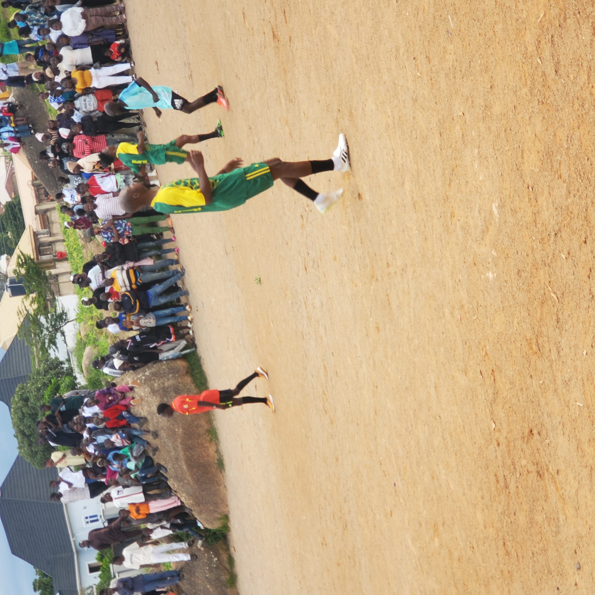 The image depicts an outdoor scene, likely a casual football match or practice, taking place on a dusty, uneven field in Jos, Nigeria. In the foreground, three young men are actively engaged in the game. One, wearing a green and yellow jersey and shorts, is in mid-stride, appearing to be dribbling or preparing to kick the ball. Another player, in a light blue jersey and black shorts, is also in motion nearby. A third individual, clad in an orange and black jersey and shorts, stands to the left of the frame, observing the action.
A crowd of spectators is gathered in the background, perched on and around large rocks and a slight incline. They are dressed in a variety of casual clothing. Behind the spectators, residential buildings with dark grey roofs are visible, along with trees and a cloudy sky. The environment suggests a typical urban or semi-urban setting in Jos, where such informal sporting events are common. The ground surface is predominantly sand and dirt, indicating a well-used playing field. The overall atmosphere appears relaxed and communal.