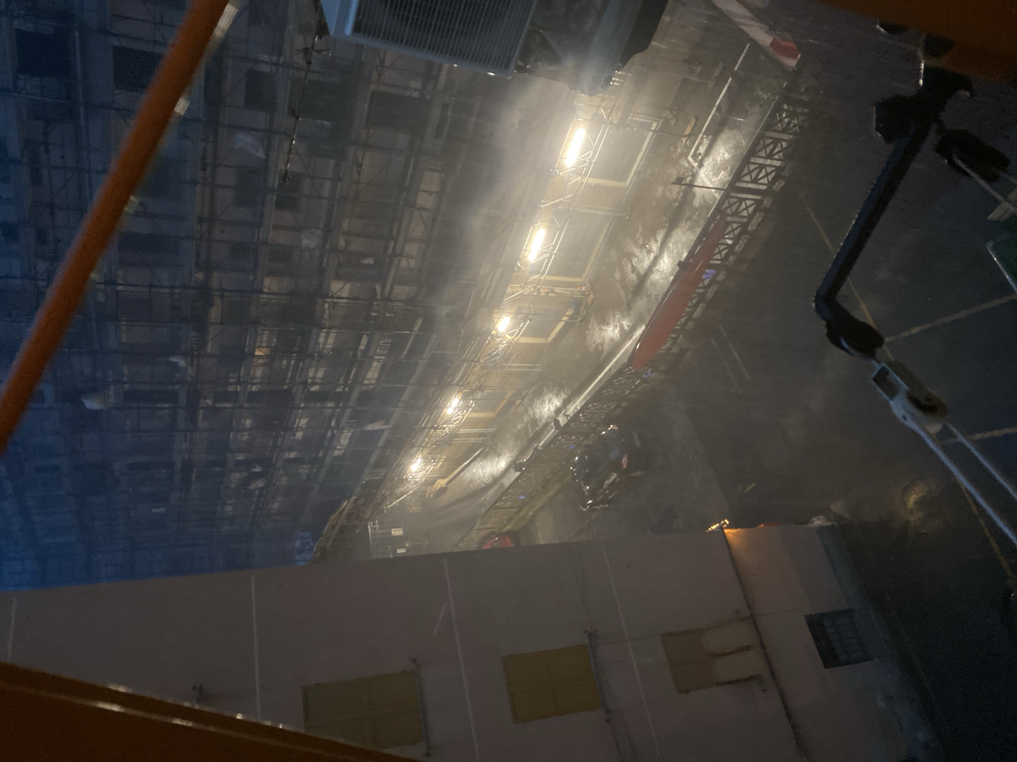 The image depicts a nighttime scene in Genoa, Italy, characterized by rain and low visibility. In the foreground, a building with light-colored walls and yellow-tinted windows is visible on the left. To the right, a street or walkway is illuminated by several bright lights, creating a hazy effect due to the rain.
The street is lined with scaffolding and construction elements, suggesting ongoing work on the buildings. A dark-colored car is parked on the street, and another red car is partially visible further down. A red barrier or banner runs along the edge of the walkway. In the background, the facade of a large building is obscured by scaffolding and the misty atmosphere. An air conditioning unit is mounted on the exterior of a building on the far right. The ground appears wet, reflecting the lights and adding to the atmospheric conditions. The overall impression is one of a wet, dimly lit urban environment at night.