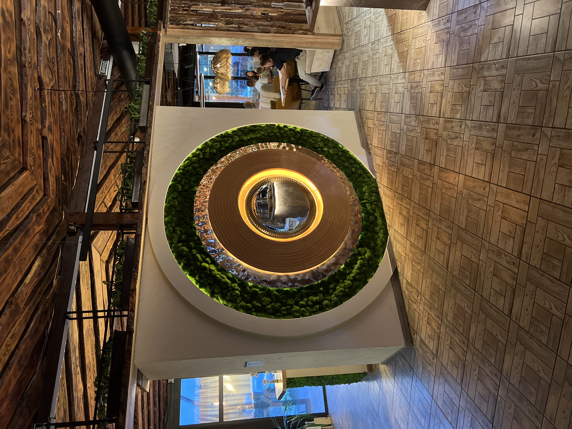 The image depicts the interior of a restaurant in Odesa, Ukraine, with a focus on a unique circular decorative element on a wall. This element features concentric rings, with the outermost ring covered in vibrant green moss, followed by a textured metallic ring, and an inner ring with a wooden finish. A mirror is at the very center of this design, reflecting some of the restaurant's interior. The ceiling is constructed of dark wooden beams and planks, adding a rustic aesthetic. The floor is tiled with a wood-patterned design. To the left, a large window shows daylight and the exterior, and a counter covered in green moss is visible. To the right, the restaurant continues with tables and patrons, including a man sitting at a table and other diners in the background. A sign with Cyrillic text, "ВИХІД" (Exit), is visible on the ceiling area.
