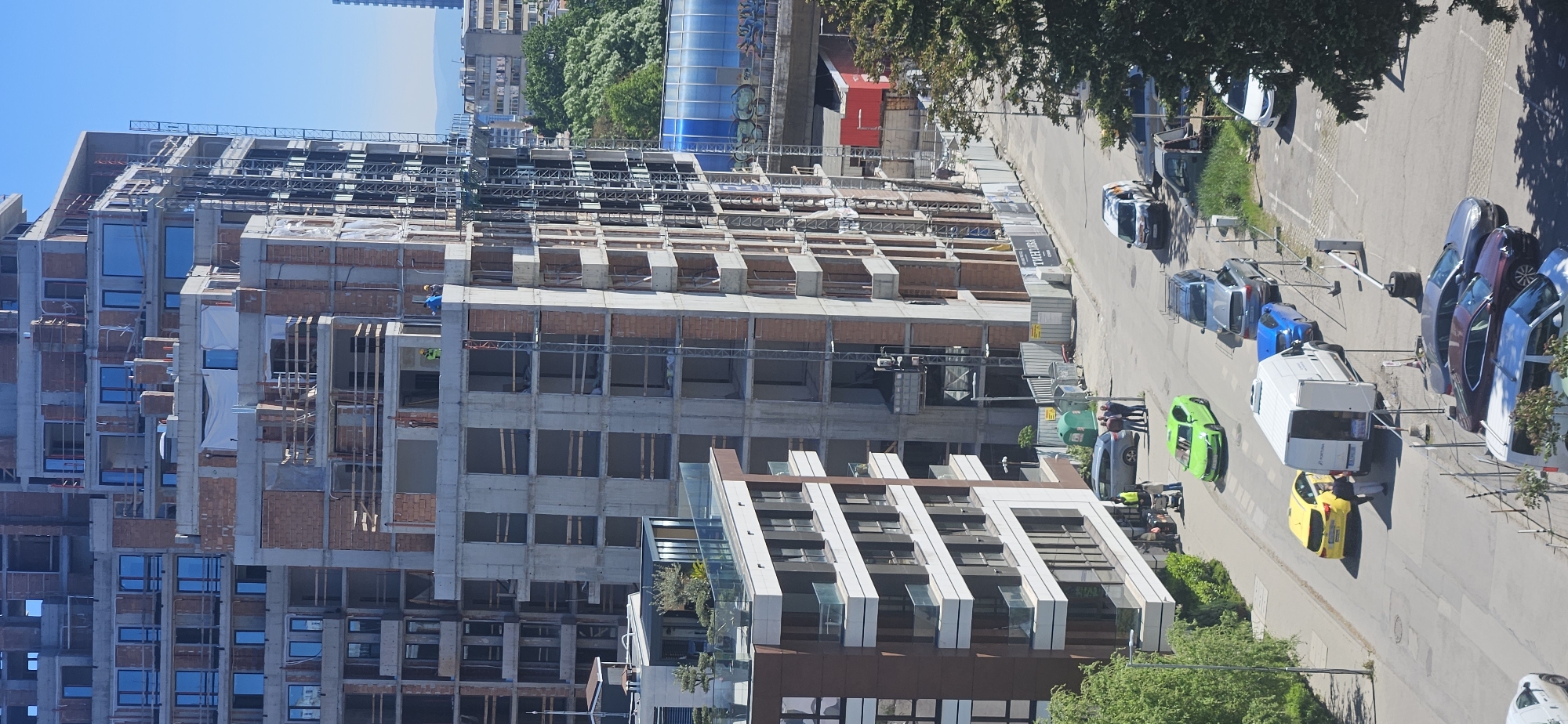 The image shows a street scene in Sofia, Bulgaria with several buildings, including a large construction site. The construction appears to be for a high-rise residential or commercial building, with partially completed brick and concrete structures. There are workers visible on the site.
A street runs alongside the building site, with various cars parked and moving along it. Car colors include yellow, green, blue, grey, white, and red. There are people interacting near the parked cars, possibly involved in loading or unloading items from a white van.
The weather is sunny, and there are trees and greenery surrounding the street. In the background, other buildings can be seen, suggesting the location is in a densely populated area.