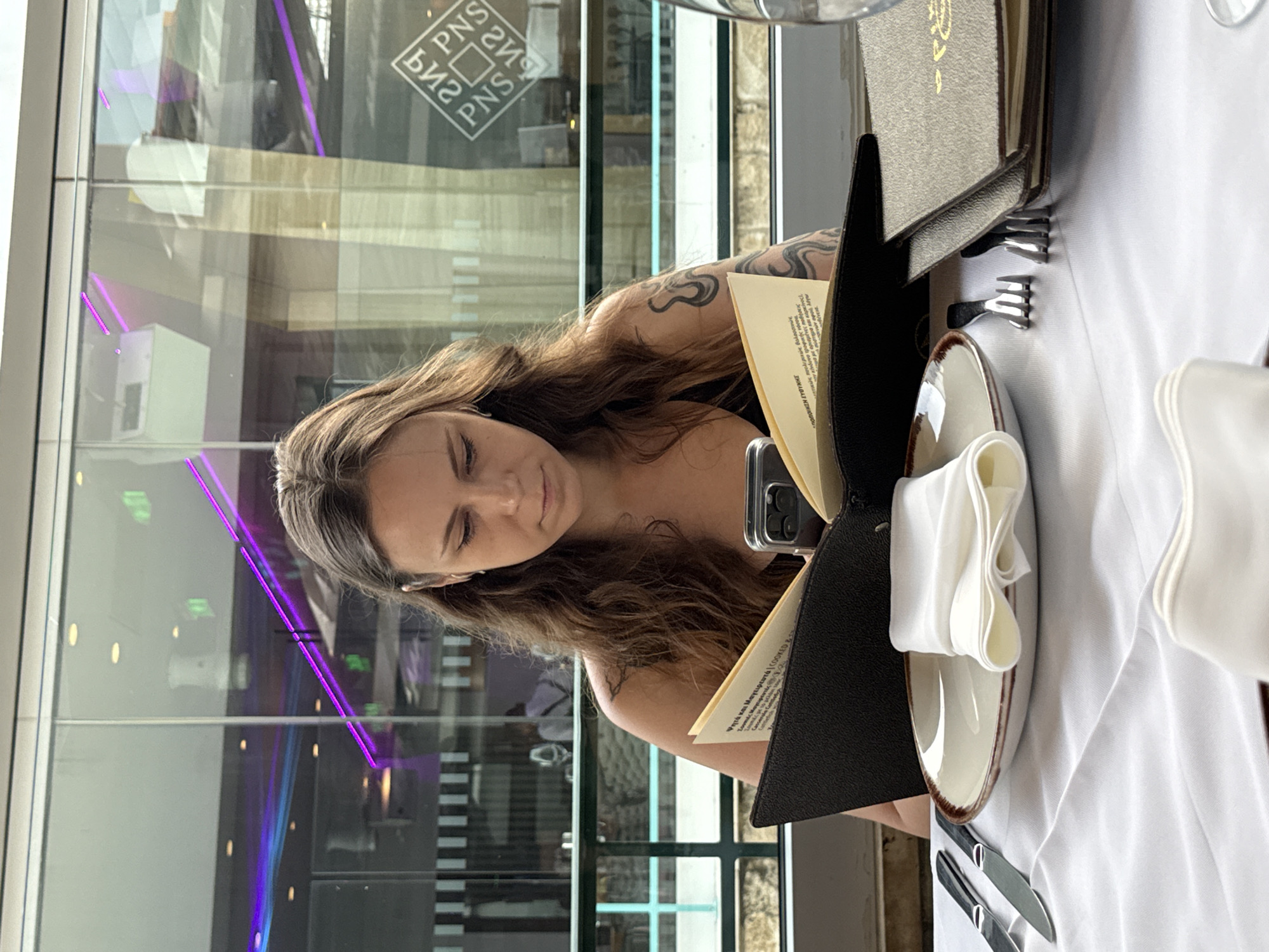 A young woman with long, wavy brown hair sits at a table covered with a white tablecloth, looking down at an open menu. She is wearing a black tank top and has tattoos on her arm. A smartphone is propped up on the menu. The table is set with a plate, a folded white napkin, and silverware. In the background, there is a glass-enclosed area with purple lighting and reflections of buildings and lights. The scene appears to be a restaurant in Limassol, Cyprus, possibly on a waterfront promenade given the reflections in the glass.