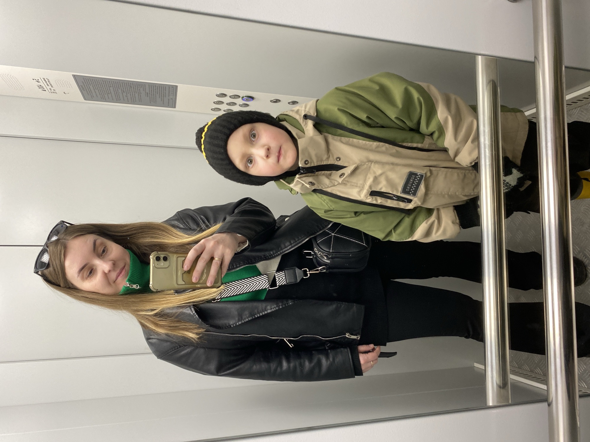 An adult female and a young male child are depicted inside an elevator cabin in Chaikovsky, Russia. The elevator's control panel, visible on the right wall, contains Cyrillic text, including "Производитель: ООО 'Пермь Монолит' Г. Чайковский ул. К. Маркса 27/1", identifying the location and manufacturer. The cabin features light gray walls, a textured metallic floor, and a horizontal chrome handrail.
The adult female, positioned on the left, has long light brown hair and black sunglasses perched on her head. She wears a black faux-leather jacket, a bright green high-collar top, a black long-sleeved undergarment, black pants, and dark footwear. She holds a gold-colored smartphone in her right hand, angled for a mirror selfie, and has a small black cross-body bag with a black and white chevron strap over her left shoulder. She looks down at the phone screen with a slight smile.
To her right stands a young male child wearing a black knit beanie with a yellow stripe and a two-toned winter jacket, beige on the upper portion and olive green on the lower portion, with visible zippers and a label on the chest. He wears black pants, with yellow footwear partially visible below the handrail. The child looks directly forward with a wide-eyed, alert expression. The scene is evenly lit, suggesting an overhead light source.
