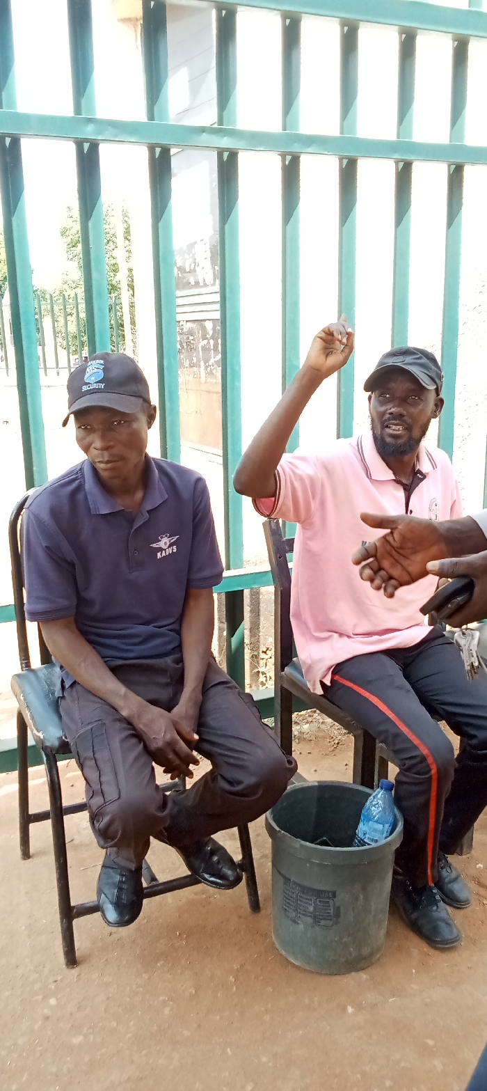 Two men are seated on simple black chairs outdoors, positioned in front of a green metal gate or fence. The man on the left wears a dark blue polo shirt with a winged "KADVS" logo, dark cargo pants, and shiny black shoes. He also wears a black baseball cap with "SHABIA SECURITY" text on the front. He is looking towards the right of the frame, with his hands clasped together in his lap.
The man on the right wears a pink polo shirt, dark pants with a red stripe down the outer seam, and a black baseball cap. He has a beard and is actively gesturing with his right arm raised, pointing upwards with his index finger, indicating he is engaged in conversation. A third person's hand is partially visible on the far right, holding what appears to be a mobile phone.
Between the two men, on the dusty ground, rests a dark grey bucket containing a blue plastic water bottle. The scene is brightly lit, suggesting it is daytime with clear weather. The attire of the man on the left, along with the gate, suggests a security checkpoint or a guarded entrance as the setting.