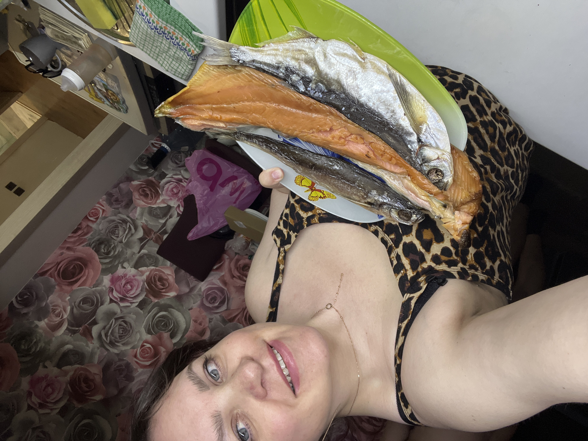 A woman with fair skin, light eyes, and brown hair is holding a stack of plates containing several dried fish. She is wearing a leopard-print top and a gold chain necklace. The location appears to be indoors. The background includes a floral-patterned floor, kitchen counter with items such as a bottle and kitchen towel, and a pink plastic bag. There's no mention of Noviy, Russia.