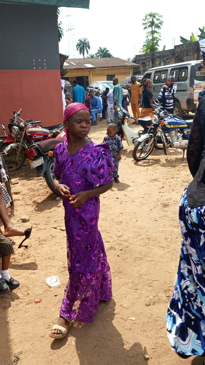 This outdoor photograph, taken in Agburu Uke, Nigeria, depicts a daytime social gathering on unpaved, sandy ground. The central figure in the foreground is a young girl, dressed in a vibrant, floor-length purple gown adorned with sequins and a matching headwrap. She wears light-colored sandals and has a neutral expression, looking to her right. Partially visible to her left are another person's feet in black shoes, and to her right, a person in a blue and white patterned skirt.
In the midground, several motorcycles are parked, including a red Qlink motorcycle behind the girl with a partially visible "ABIA" license plate, and a blue Qlink motorcycle further to the right. A small child in a patterned blue outfit stands behind the girl. Beyond these, a group of adults and children are gathered, some seated on benches, others standing and conversing. Their attire ranges from casual to semi-formal, including traditional wear like an orange outfit on one man.
The background features various vehicles, including a white van with partially visible text "SOLAS AS MOTOR..." and "UMTC". Simple buildings with corrugated iron roofs are visible under a bright, sunny sky. Palm trees and other vegetation dot the landscape, characteristic of a rural or semi-urban African setting. The presence of strong shadows suggests a clear, bright day. The overall atmosphere is one of a calm community gathering.