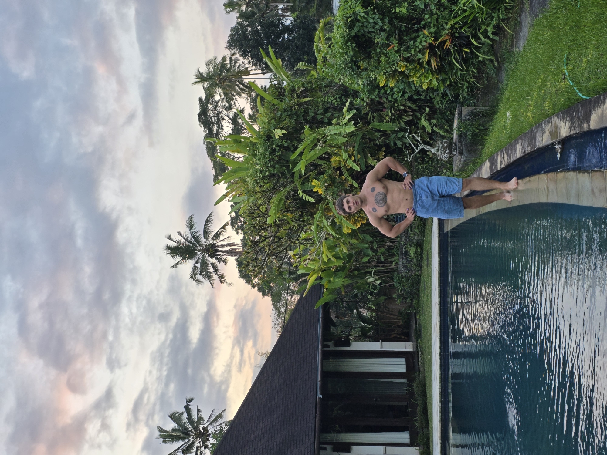 The image captures a shirtless man standing at the edge of a swimming pool in a lush, tropical setting. He is wearing blue shorts and has tattoos on his chest and torso, as well as a watch on his left wrist. His bare feet are partially submerged in the dark blue pool water, which reflects the sky and surrounding greenery.
The scene is dominated by dense, verdant foliage, including large banana-like leaves and several tall palm trees, which frame the background. Behind the man, an abundance of green plants creates a vibrant, natural backdrop. To the right of the pool, a section of green grass with a light green garden hose is visible.
In the lower left corner, a portion of a building with a dark, sloped roof and several large windows can be seen, partially obscured by more palm trees. The sky above is partly cloudy with soft, pastel shades of pink and orange peeking through the grey clouds, suggesting either a late afternoon or early morning light. The overall atmosphere is serene and tropical. There is no visible text in the image.