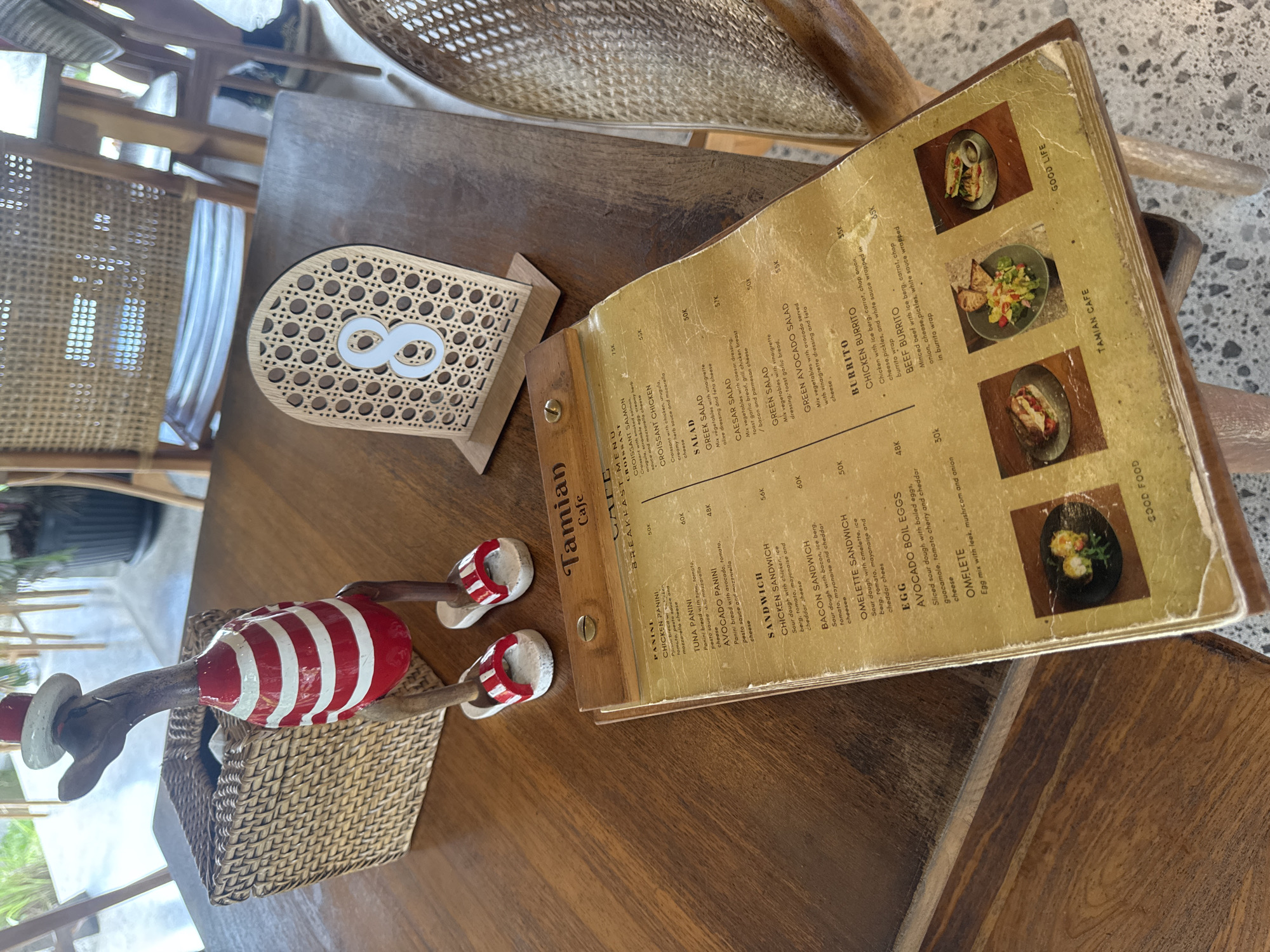 A wooden table in the city of Tegallalang, Indonesia, is set with a menu, a decorative wooden duck figurine wearing a red and white striped shirt and a red hat, and a small woven basket. The menu, which is open, displays various food items. A table number sign with the numeral '8' in white is positioned on the table. The environment suggests an outdoor seating area of a cafe, with glimpses of chairs and plants in the background.