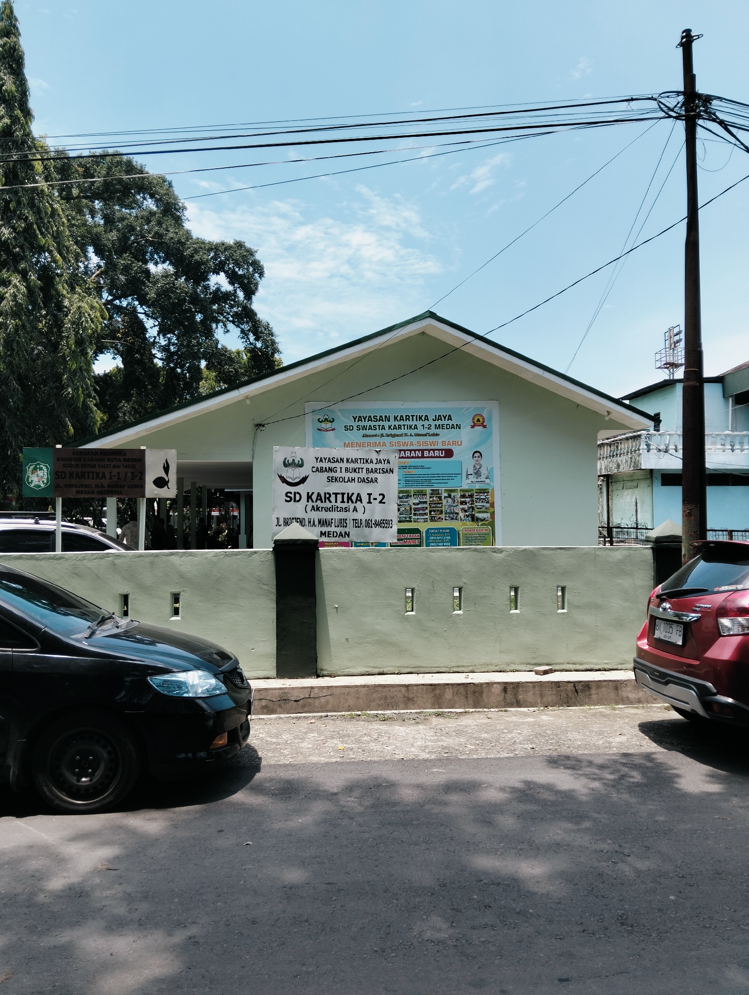 This is a medium shot, eye-level, outdoor photograph taken on a sunny day in the city of Medan, Indonesia. The scene is a street in front of a light green, single-story building with a dark green roof. The building appears to be a school, as evidenced by signs displaying "SD KARTIKA I-I / I-2" and "SEKOLAH DASAR". One large banner on the building reads "YAYASAN KARTIKA JAYA SD SWASTA KARTIKA 1-2 MEDAN MENERIMA SISWA-SISWI BARU TAHUN AJARAN BARU".
Two cars are parked on the asphalt road in front of the school. A black car is parked on the left side of the image, facing right, with its front end visible. A red car is parked on the right side of the image, facing left, with its rear end visible. The red car has a license plate that reads "BK 1035 FB".
A low, light green concrete wall with square cutouts runs along the front of the school property. Several trees with dark green foliage are visible behind and to the left of the building. A utility pole with numerous wires is situated on the right side of the image, behind the red car. The sky is blue with scattered white clouds. There are no people visible in the image.