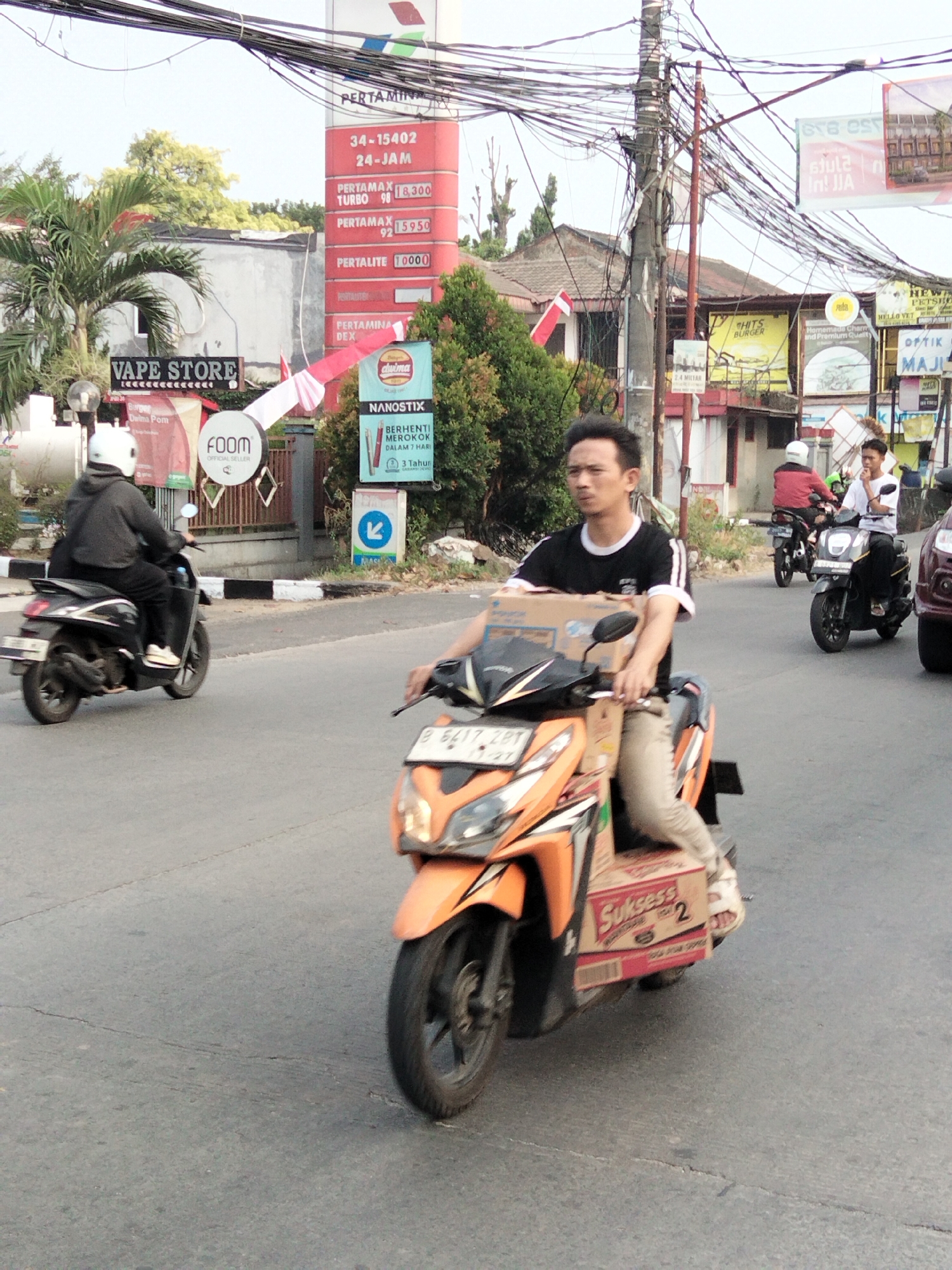 This is a street-level, medium-distance, eye-level shot of a man riding an orange and black scooter, carrying a box labeled "Sukses" on the back. The man is wearing a black t-shirt with white stripes on the sleeves, light-colored pants, and white sneakers. He is looking slightly to his left. The scooter is in the foreground, with its front wheel turned slightly. In the background, another person on a scooter, wearing a white helmet and a gray hoodie, is also visible. To the right, another scooter with a red top rider and a white helmet is further down the road. Behind the scooters, there are buildings, including a gas station with prices listed for various fuels like PERTAMAX TURBO, PERTAMAX, and PERTALITE. There are also various signs, including "VAPE STORE" and "FOOM". The scene appears to be in a busy urban environment, likely in South Tangerang, Indonesia, indicated by the presence of the Pertamina gas station branding and common street scenes in Indonesia. The road is paved and shows signs of wear. Overhead power lines crisscross the sky, a common sight in Indonesian cities. The overall lighting suggests a daytime setting, likely under a bright sky with some haze.