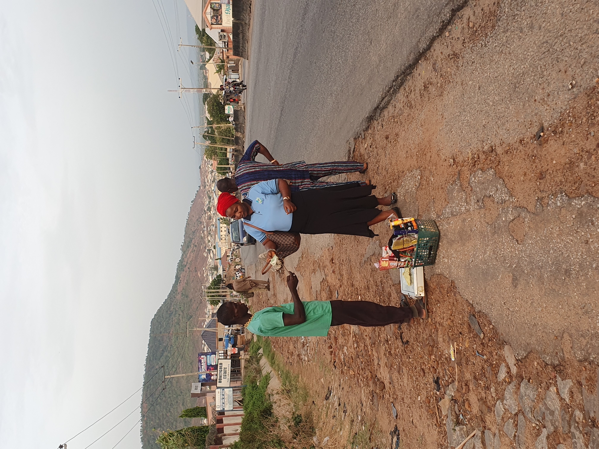 The image depicts a scene on a roadside in Lokoja, Nigeria. A man in a green t-shirt and dark trousers stands facing a woman in a blue shirt, black skirt, and red head covering. She holds a brown handbag and is engaged in what appears to be a transaction with the man. Beside the woman, a man in a patterned blue and brown outfit stands with his hand on his hip. A green basket filled with various items, including snacks and other packaged goods, sits on the ground near the woman. The roadside is unpaved and dusty. In the background, the landscape includes buildings, trees, power lines, and a hill. Roadside signage indicates "Honeygold Estate" and other advertising. A vehicle is visible on the road behind the woman. The weather appears overcast.