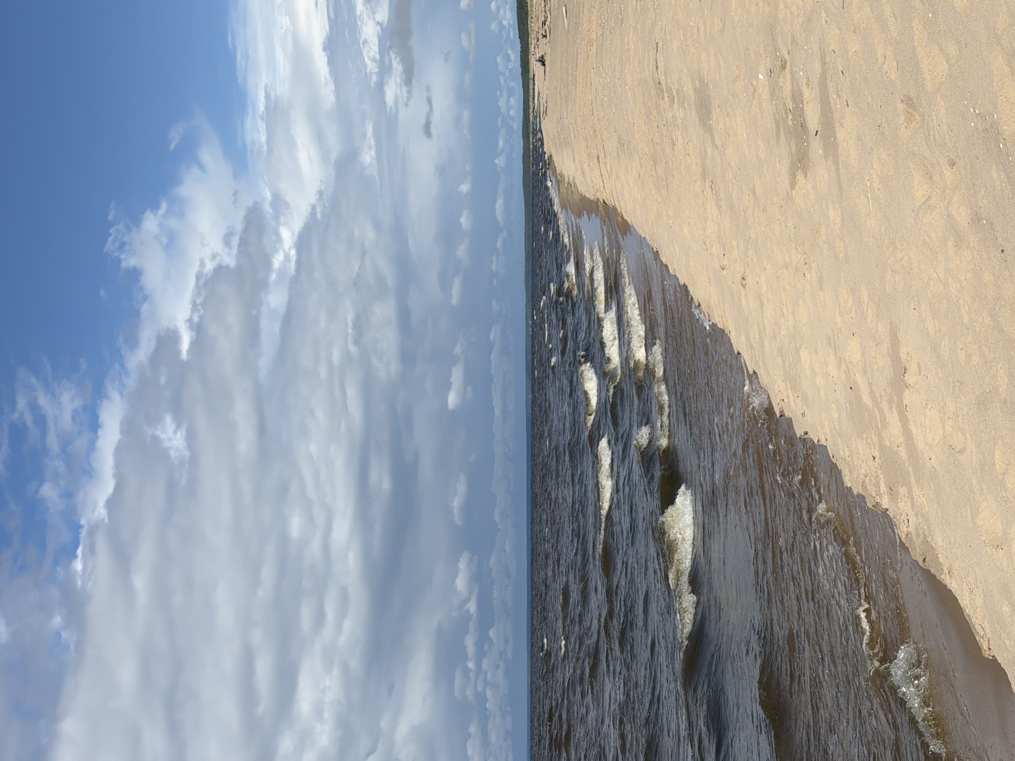 This is a full shot photograph of a beach scene on a cloudy day. The lower left quadrant shows dark, choppy water with white-capped waves rolling towards a sandy shore. The beach occupies the right half of the frame, appearing tan and lightly textured with small scattered white shells. In the distance, along the coastline, a treeline is visible, and a few small figures can be seen sitting or standing on the sand. The upper portion of the image is dominated by a dramatic sky filled with heavy, grey clouds, interspersed with patches of blue sky. The location is identified as Gauja, Latvia.