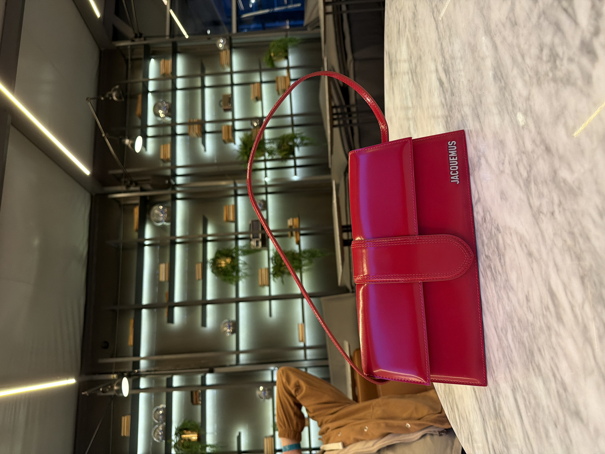 A bright, vertical indoor photograph captures a red leather Jacquemus handbag resting on a light marble surface. The bag is oriented vertically, with the “JACQUEMUS” logo visible on its right side. A section of a person is visible at the bottom of the image, wearing tan-colored pants and a light-colored jacket. The background features a wall structure with vertical bars and shelves, punctuated with foliage. Illuminated light fixtures are visible in the upper part of the image. The photo provides no direct evidence to verify its location in Krasnoyarsk, Russia.