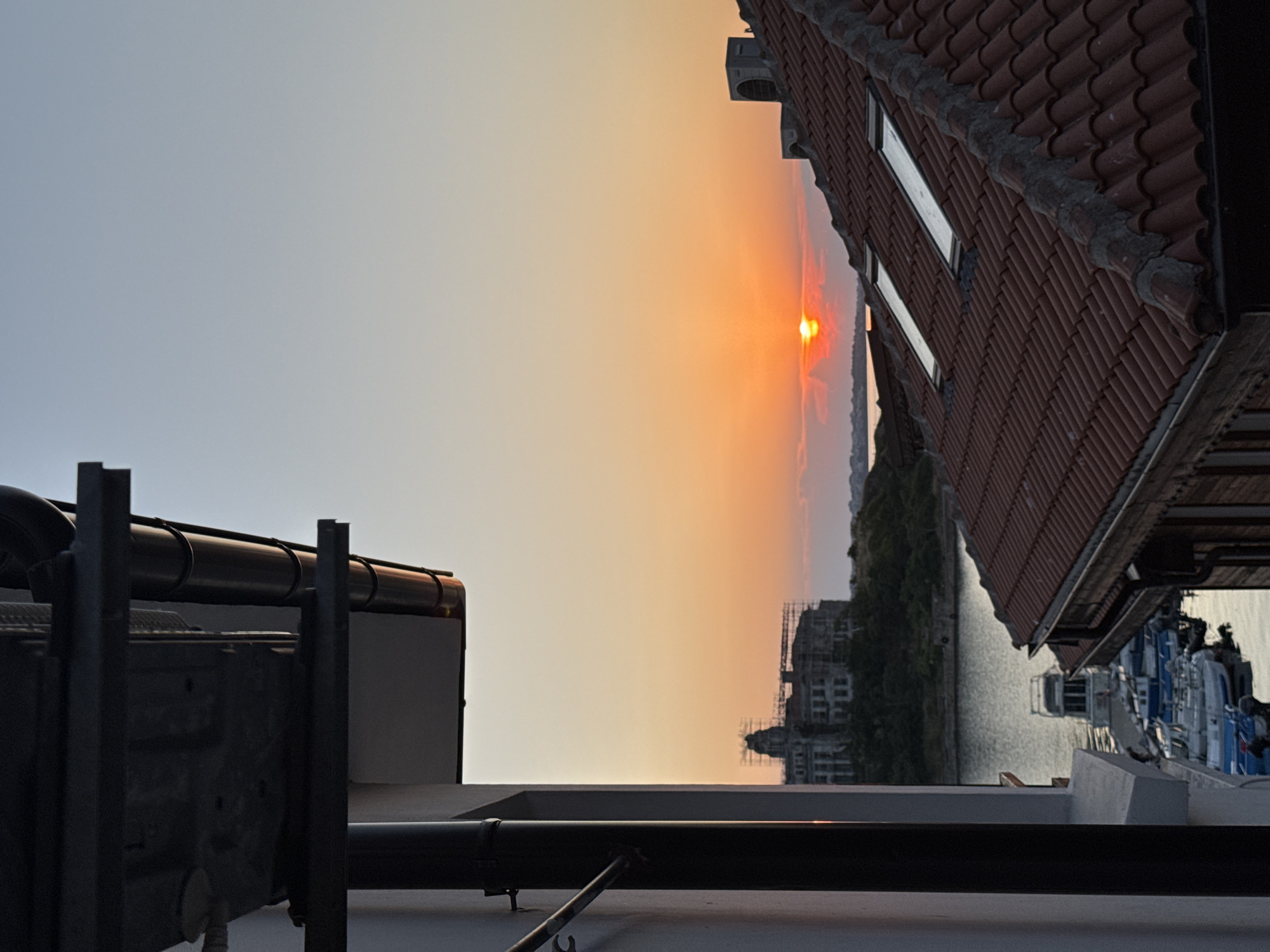The image depicts a sunset scene in Sozopol, Bulgaria. The sun is low on the horizon, casting a warm orange glow across the sky. The foreground features rooftop elements, including pipes, railings, and part of a building with a tiled roof. To the right, a building with a red-tiled roof and windows is visible. In the background, across a body of water, there is a tree-covered hillside and buildings, some of which appear to be under construction. Boats are docked along the waterfront on the right side of the image. The overall mood is peaceful and serene, characteristic of a sunset. The time of day is clearly evening, as indicated by the setting sun.