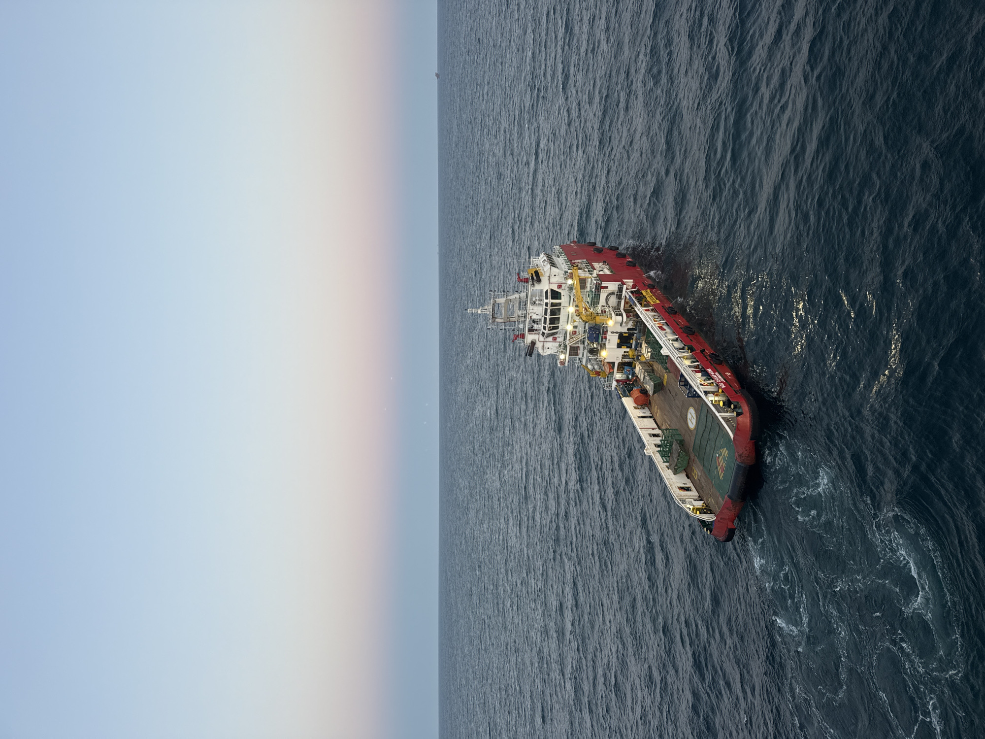 The media file shows an aerial view of a ship sailing on a dark, choppy ocean. The ship is red and white and appears to be a supply vessel, with a helideck on its stern. The ocean stretches to the horizon, meeting a pale sky that transitions from light blue to a soft yellow and pink hue, suggesting either dawn or dusk. The scene is serene and vast, with no other vessels or land in sight.
The ship's hull is visible along with the deck structures, including cranes and what appears to be cargo. The water's surface is disturbed by the ship's movement and natural waves, creating white foam and swirling patterns. The lighting indicates it's either early morning or late evening, with the sun low on the horizon casting a gentle light. The overall mood is tranquil, emphasizing the isolation of the vessel at sea.
There is visible text on the side of the ship, which appears to read "VETERAN WIRE 829". The setting is open ocean, and the weather appears calm despite the choppy water. No people are clearly visible in the image. The composition emphasizes the scale of the ocean and the solitary nature of the ship.