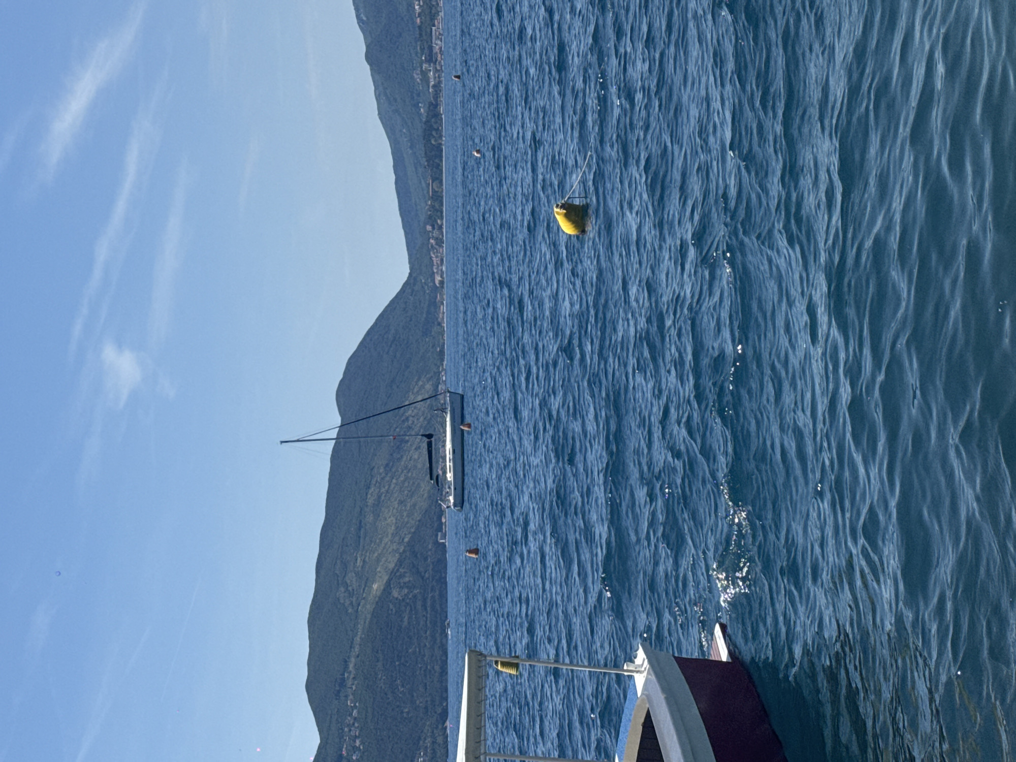 The image depicts a serene maritime scene under a clear blue sky, likely during daytime, at an Unknown location. In the foreground, the bow section of a small boat is visible on the left, featuring a maroon hull with a white trim and a white canopy structure. A small, golden bell-like object hangs from the canopy's interior. The deep blue water in front of the boat shows gentle ripples with scattered sunlight reflections.
In the midground, the expansive body of water contains several objects. A prominent yellow cylindrical buoy, secured by a white rope, floats slightly right of center. Further back and to the left, an orange cylindrical buoy is visible. Another smaller orange buoy floats even further back towards the right. A white sailboat, with its mast erect and rigging visible but sails furled, is anchored or adrift in the water towards the left of the frame.
The background is dominated by a continuous range of lush, green, tree-covered mountains stretching across the horizon. Along the base of these mountains, particularly on the right side, small clusters of buildings indicative of a coastal settlement are discernible. The sky above is a bright, clear blue, with minimal, faint wispy white clouds, primarily in the upper right quadrant. No individuals are present in the frame.
