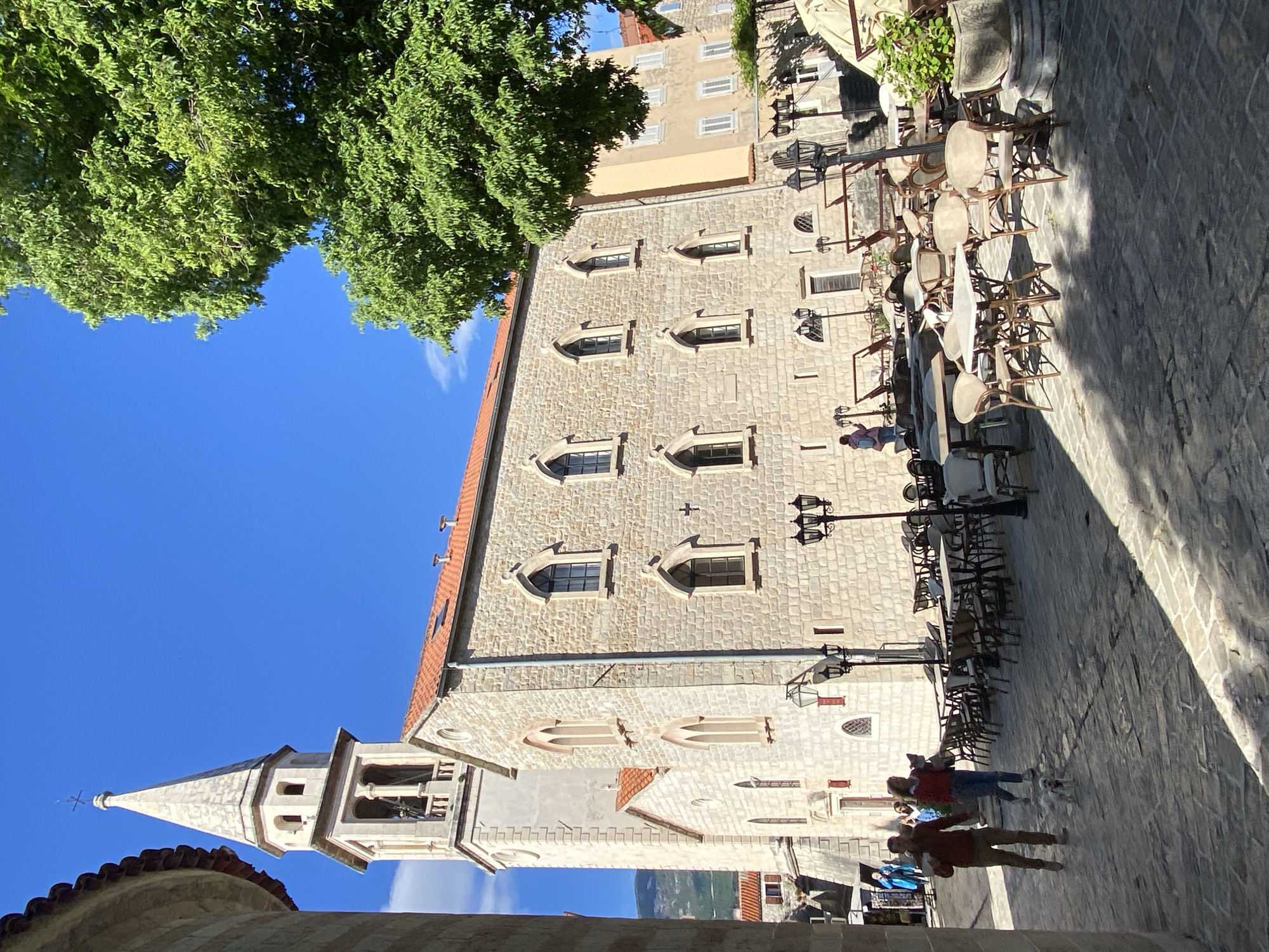 The image captures a vibrant street scene within a historic town, likely Budva, Montenegro, under a clear blue sky. A large, multi-story building constructed from rough-hewn light-colored stone dominates the left side of the frame. This building features distinctive arched windows with intricate stone detailing, arranged in several rows. Its red-tiled roof is visible at the top. To the far left, a tall church tower with a white stone spire and arched bell openings stands against the bright sky, topped by a small cross.
In the upper left corner, a dense canopy of lush green tree foliage extends into the blue sky, partially shading the area. The foreground and right side of the image show a stone-paved street or plaza, serving as an outdoor seating area for a cafe or restaurant. Numerous tables and chairs, some neatly stacked and others arranged, line the side of the stone building. Several ornate black street lamps with multiple lanterns are also present.
A few people are scattered throughout the scene. In the lower right, two individuals are walking, one of whom appears to be leading a small white dog on a leash. Another person is visible further back near the cafe seating. The scene is brightly lit by the sun, casting long, distinct shadows across the stone pavement from the buildings and street furniture, indicating either late morning or early afternoon. The weather is sunny with only a few wispy clouds visible in the sky. No discernible text is visible in the image.