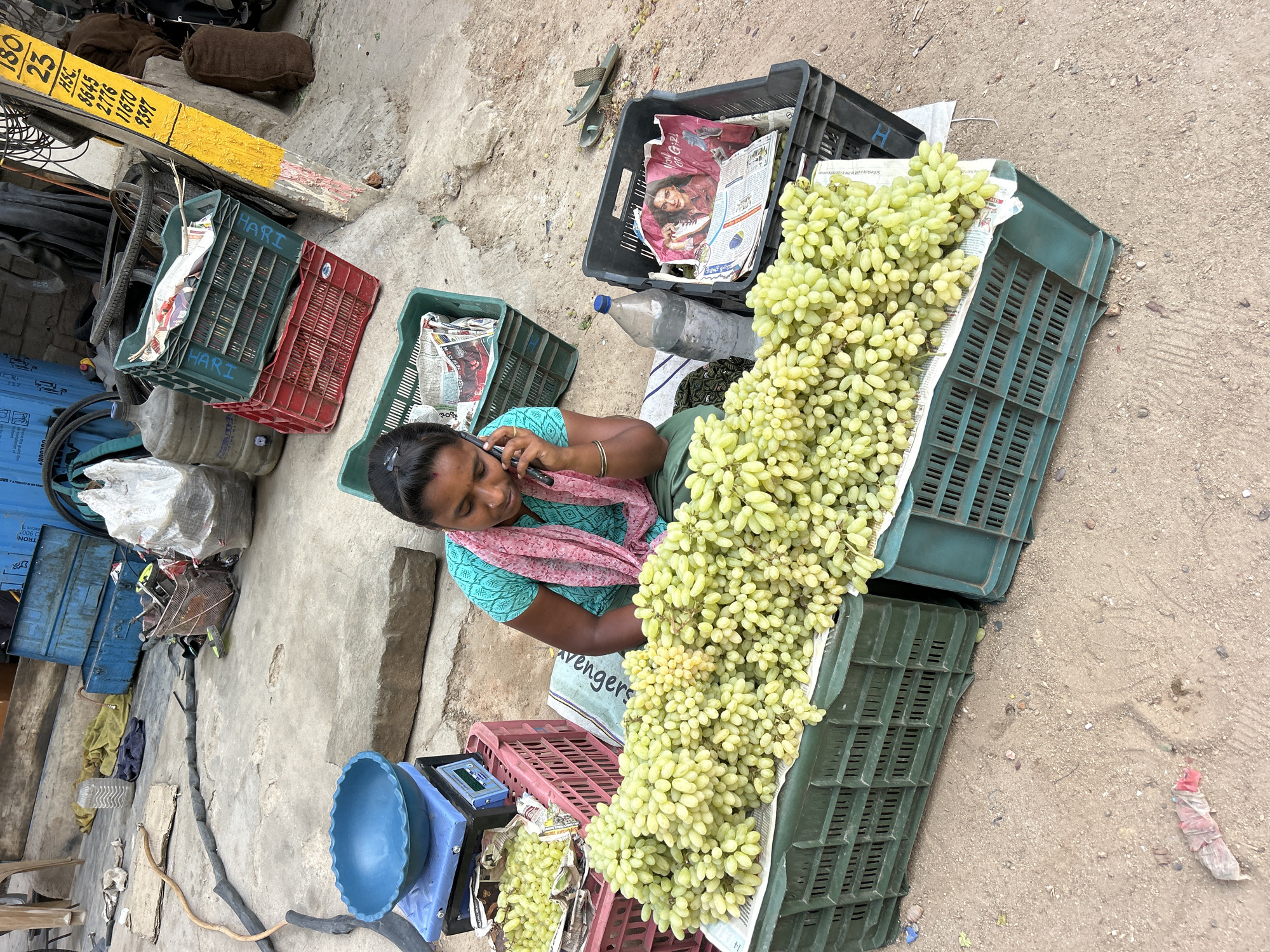 The image depicts a street scene in Puttaparthi, India. An Indian woman is seated on the ground, speaking on a cell phone, while surrounded by crates of green grapes that appear to be for sale. There are at least six crates of grapes in various states of fullness and arranged adjacent to her. A blue scale is near the woman, and newspaper is used to line some of the crates. Other items nearby include water bottles, additional crates, and street debris. The environment appears to be a sidewalk or similar paved area with a dirty surface.